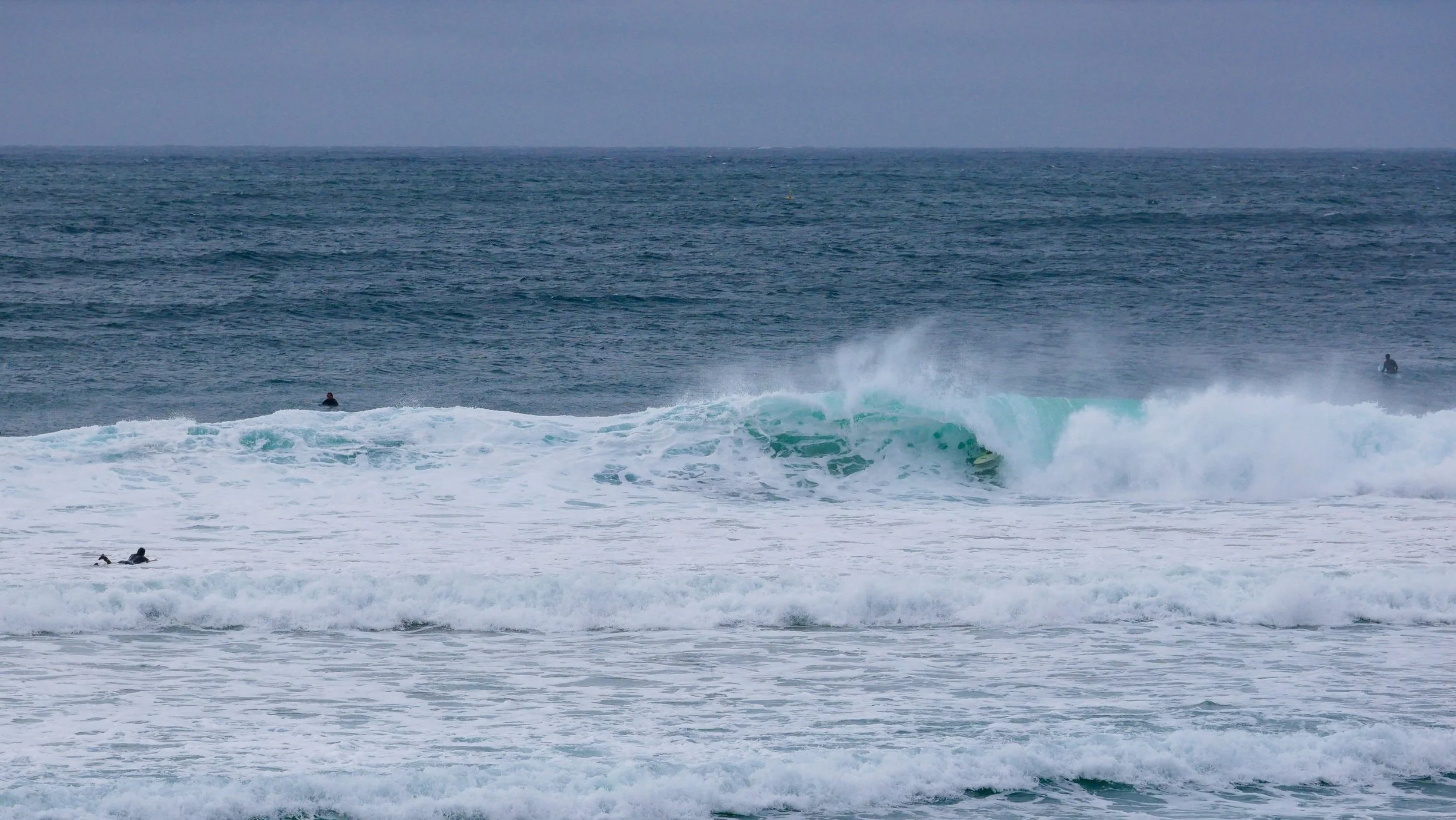 Surfers in wetsuits riding waves in the ocean under a cloudy sky.