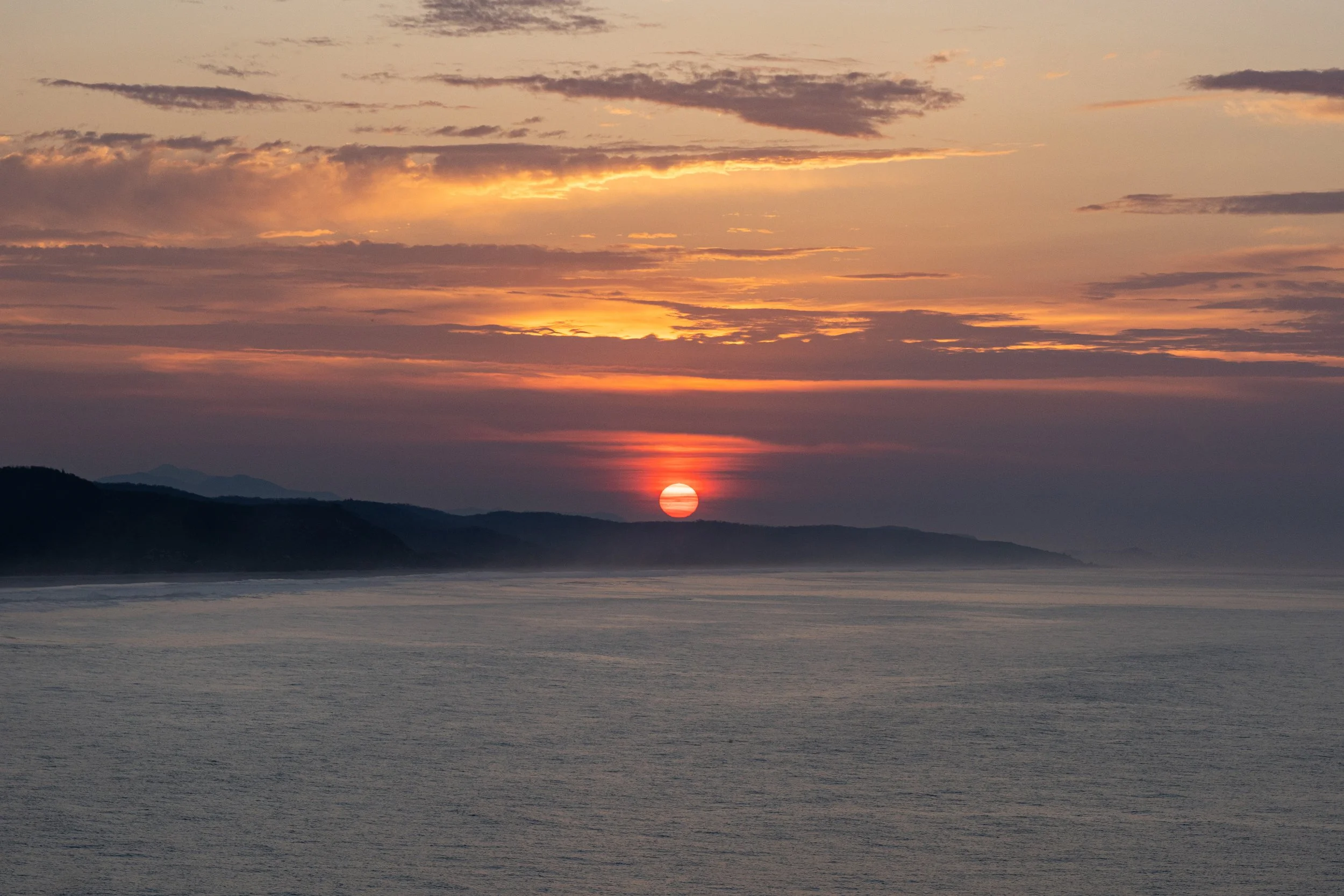 Sunset over a body of water with mountains in the background, some clouds in the sky, and the sun partially below the horizon.