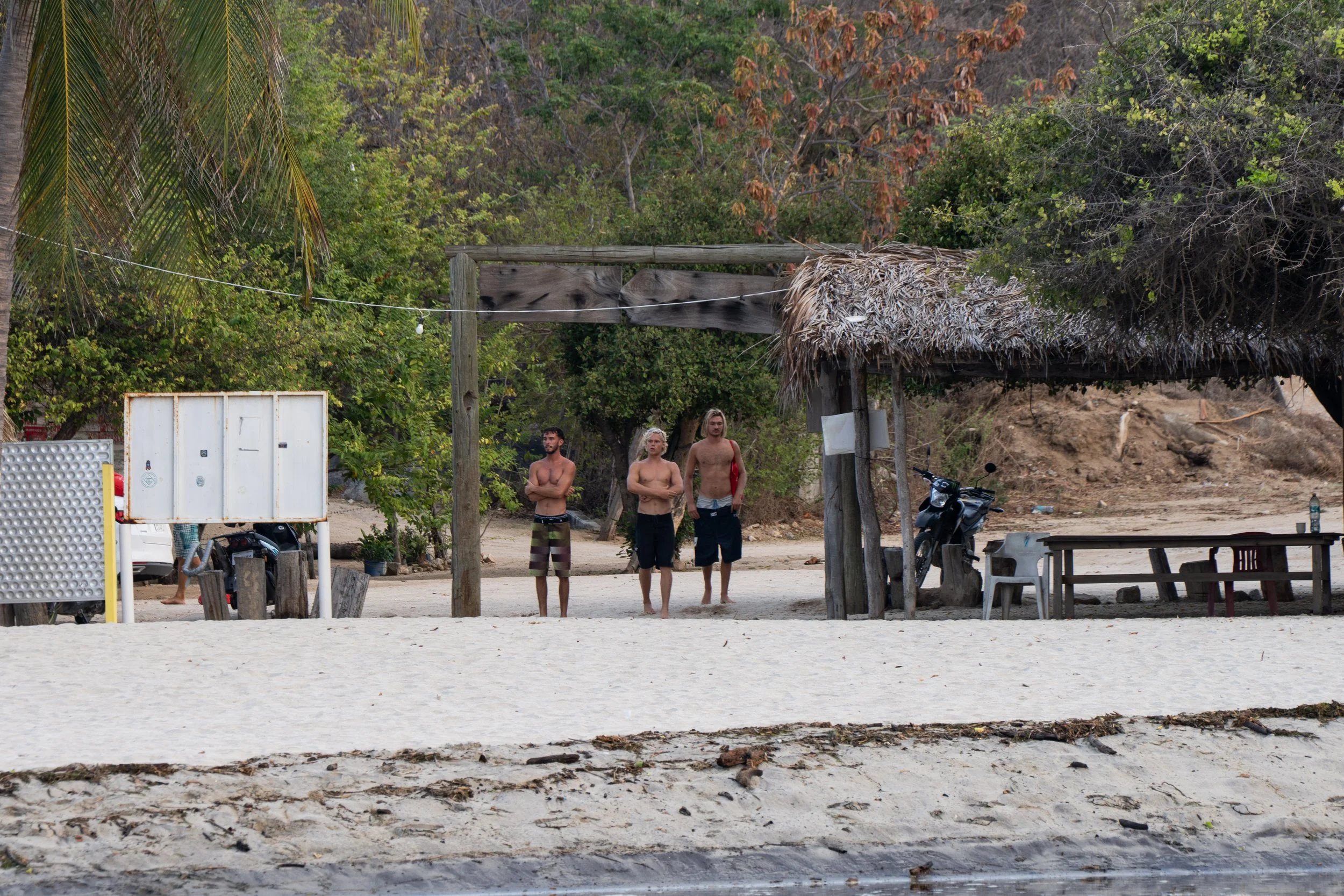 Three shirtless men standing on a sandy beach near a rustic thatched roof hut, with tropical trees and foliage in the background.