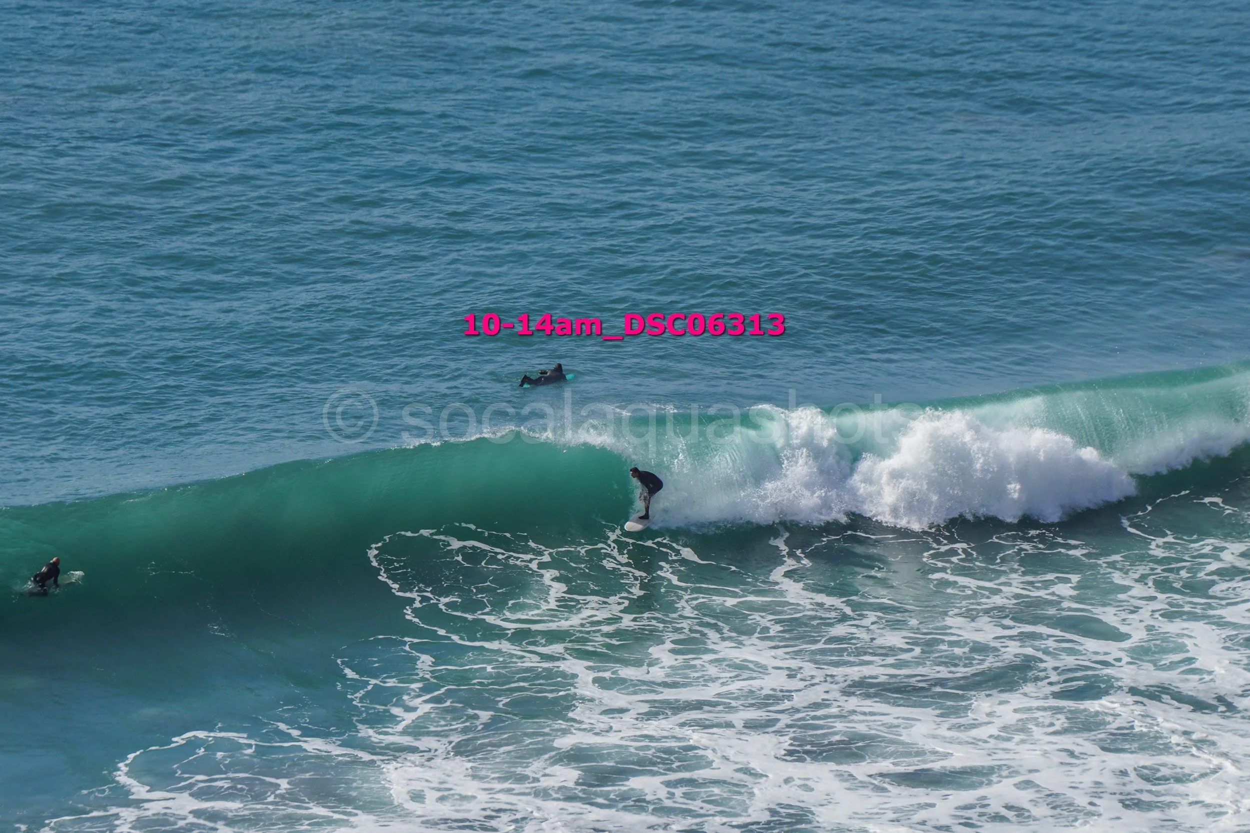 Three surfers in wetsuits riding and waiting for waves on the ocean with visible foam and white water, clear blue sky in background.