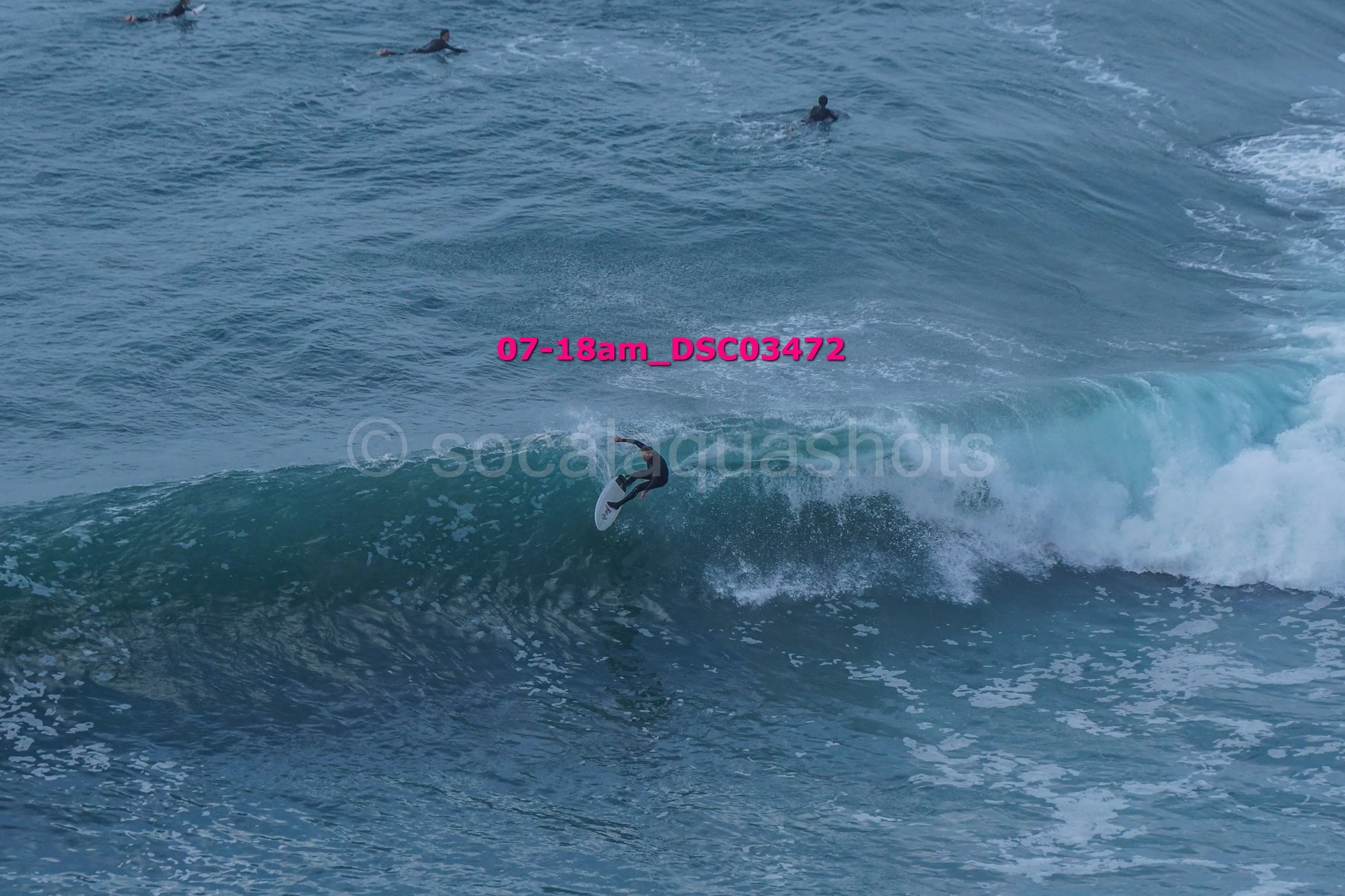 Surfer riding a wave in the ocean with several people swimming in the background.