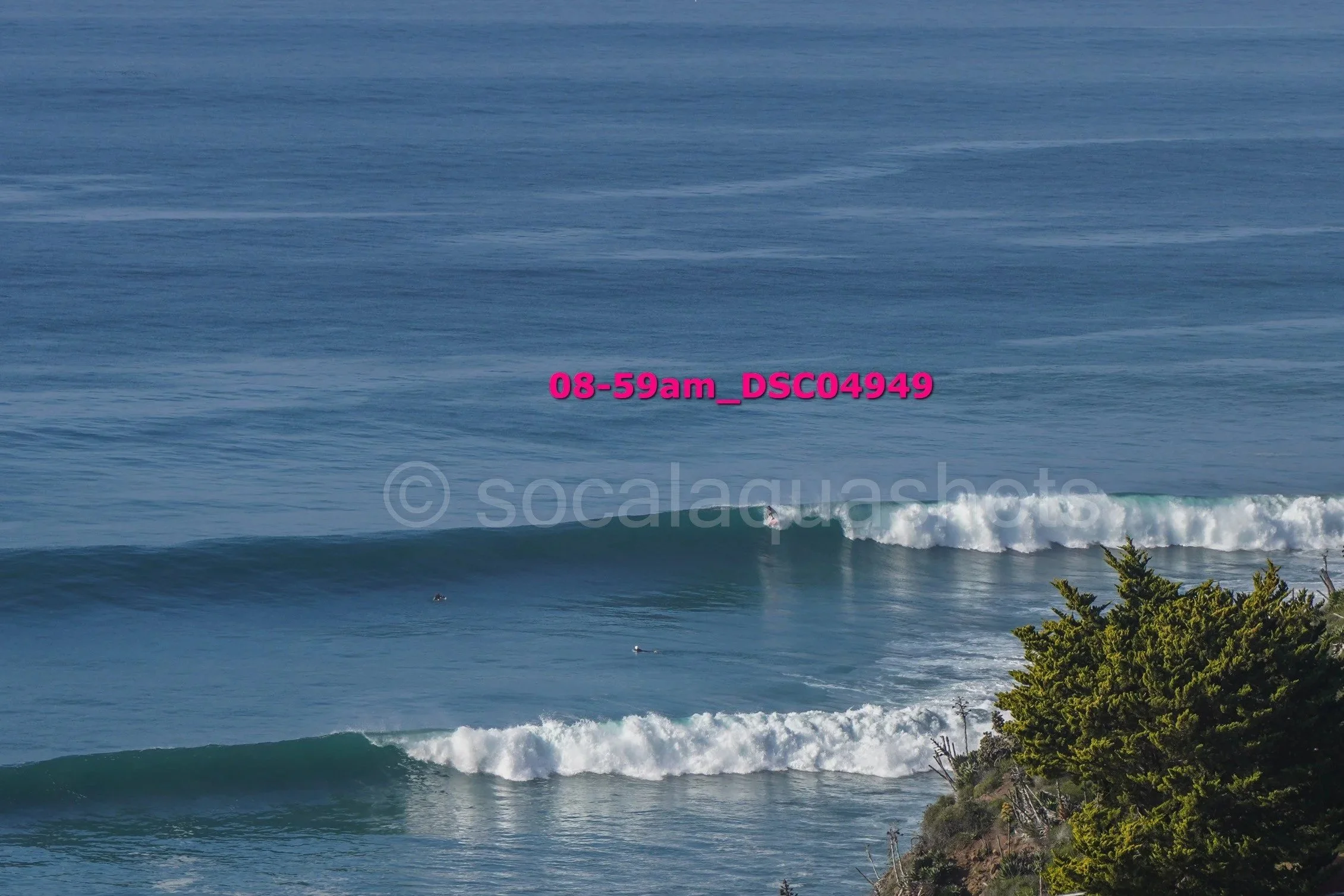 Ocean waves crashing near a rocky shoreline with green trees, blue sky, and surfers in the water.
