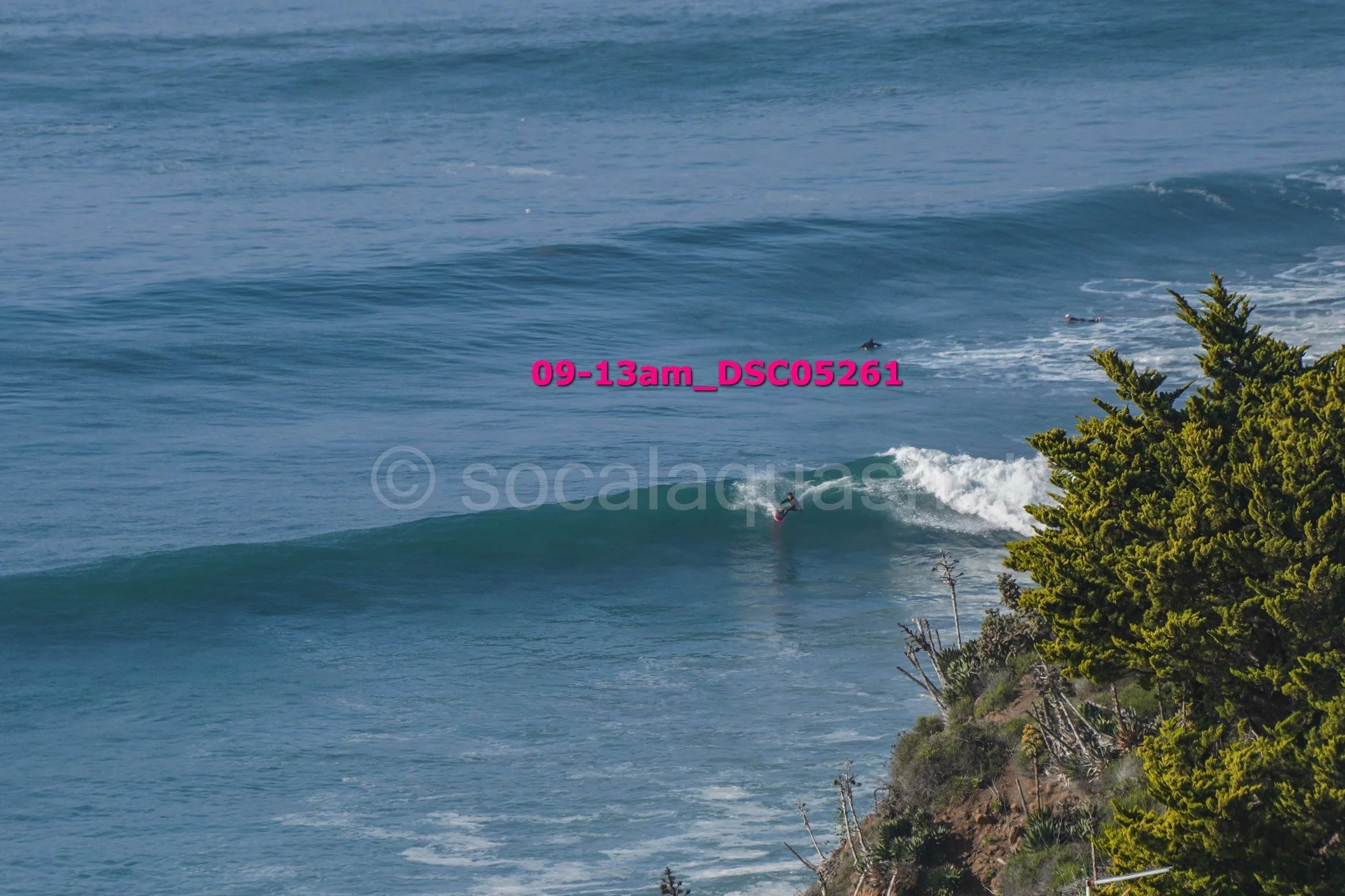 Surfer riding a wave near a rocky coastline with trees in the foreground, ocean in the background, timestamp 09-13am, water and sky.