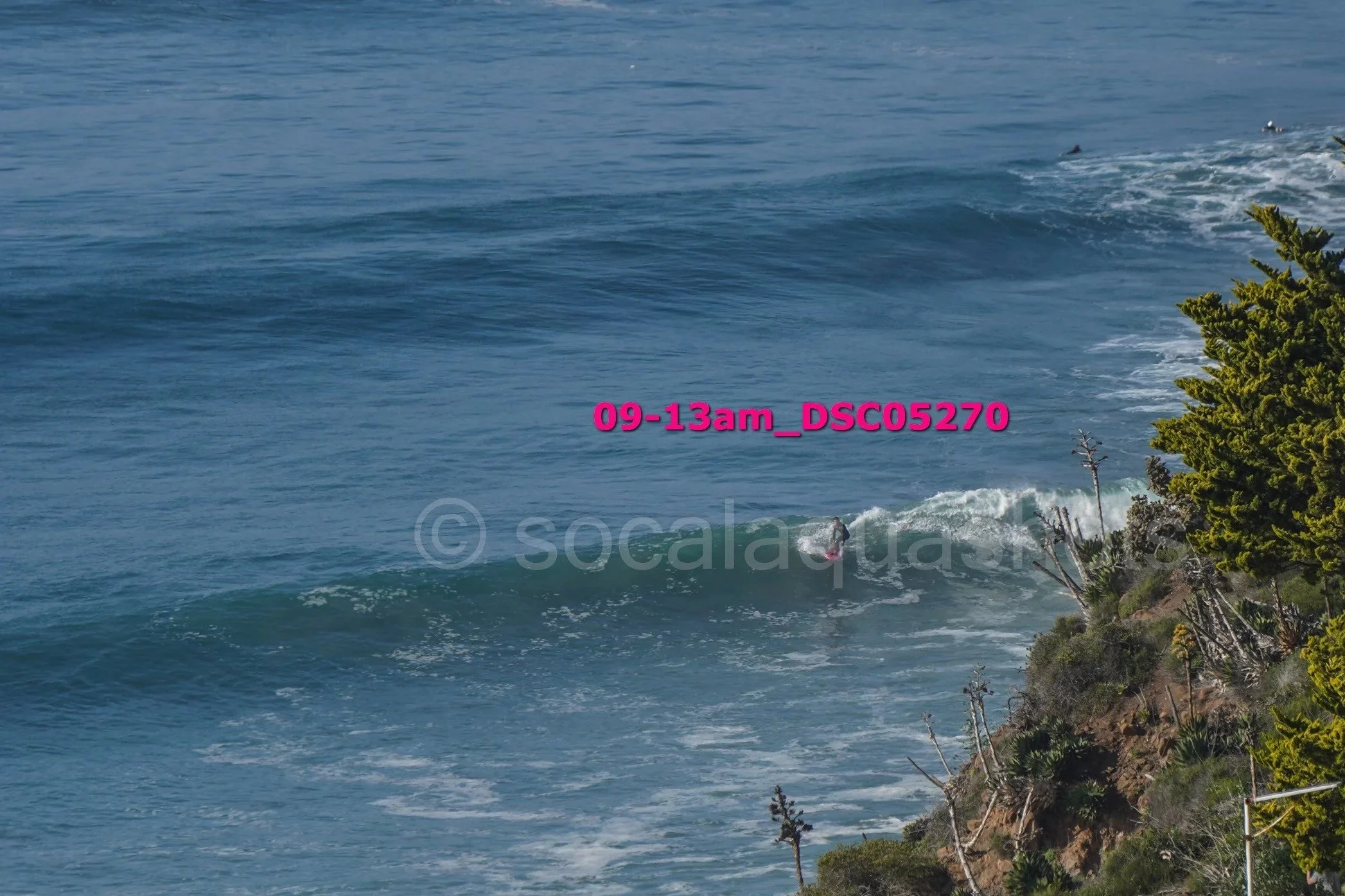 A surfer riding a wave near a rocky cliff with green bushes and trees, ocean in the background, and a timestamp overlay reading 09-13am_DS05270.
