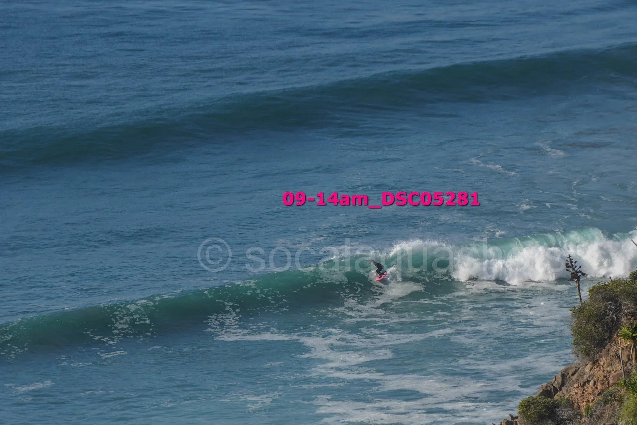 A person surfing on a small wave near a rocky shoreline with the ocean in the background.