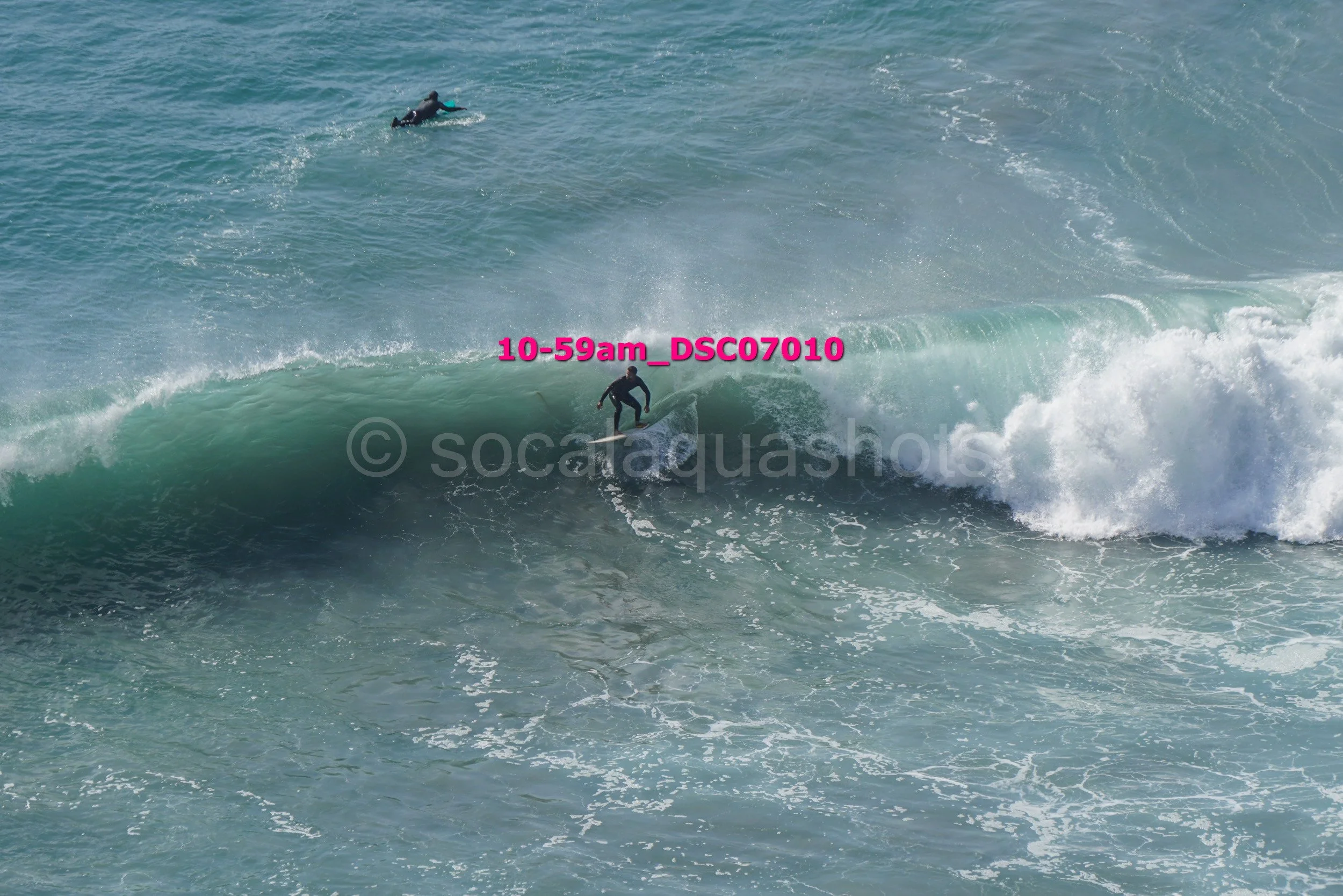 A person surfing on a large wave in the ocean, with another surfer visible in the background.