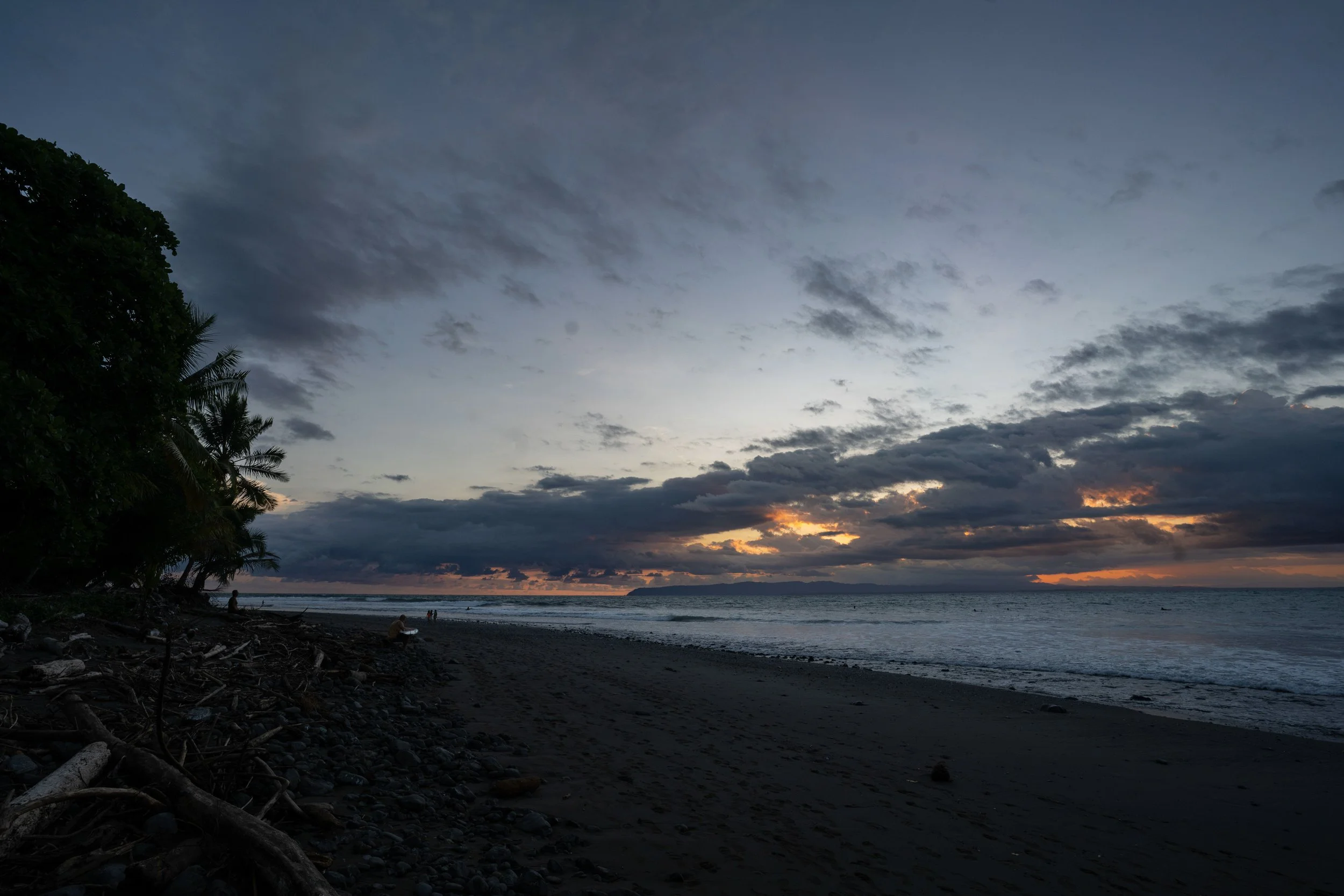 A beach scene at dusk with a dark, cloudy sky. There are some trees on the left side and a few people along the shoreline. The ocean is calm with gentle waves, and the horizon shows a faint glow of the setting sun.