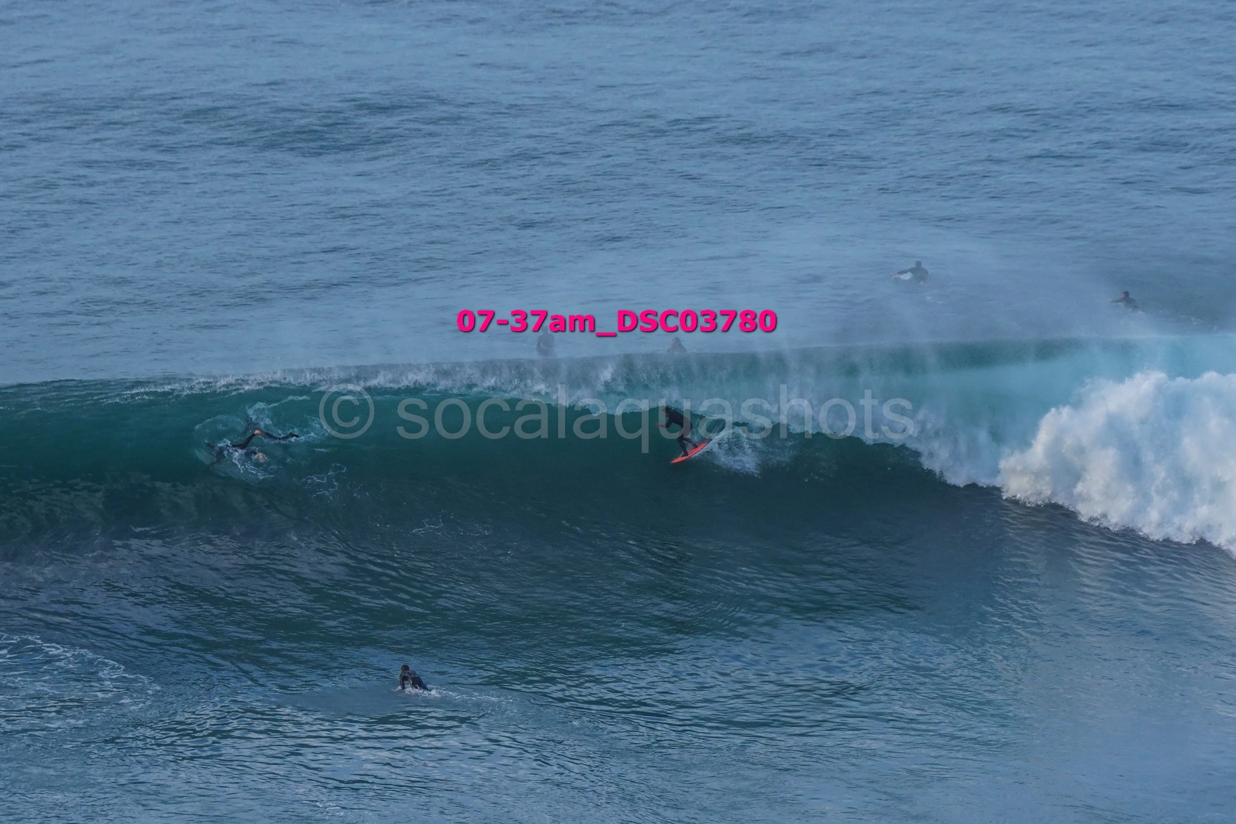 Surfers riding large ocean waves with other surfers in the background, some paddling and waiting for waves.