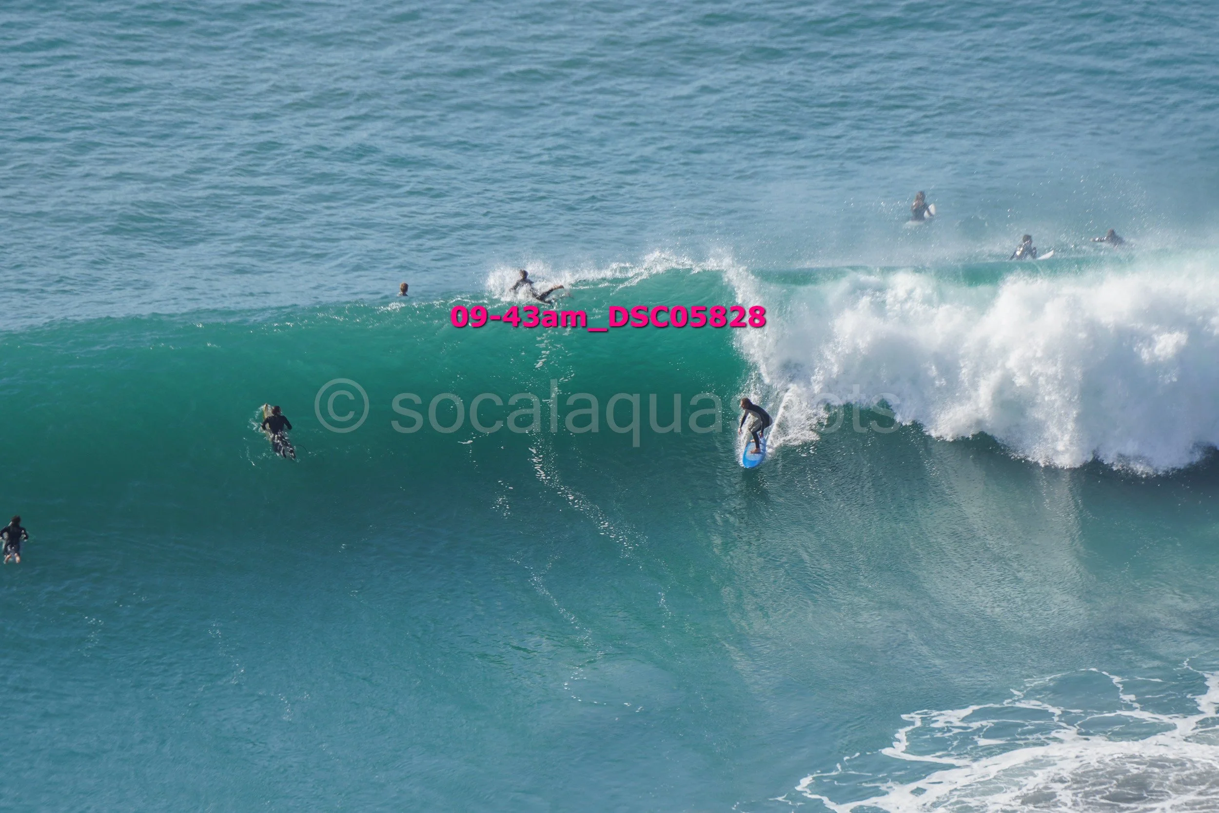 Surfers riding and waiting for waves in the ocean, with some on surfboards and others on their boards. The water is a deep blue-green, with white foam from the breaking waves.