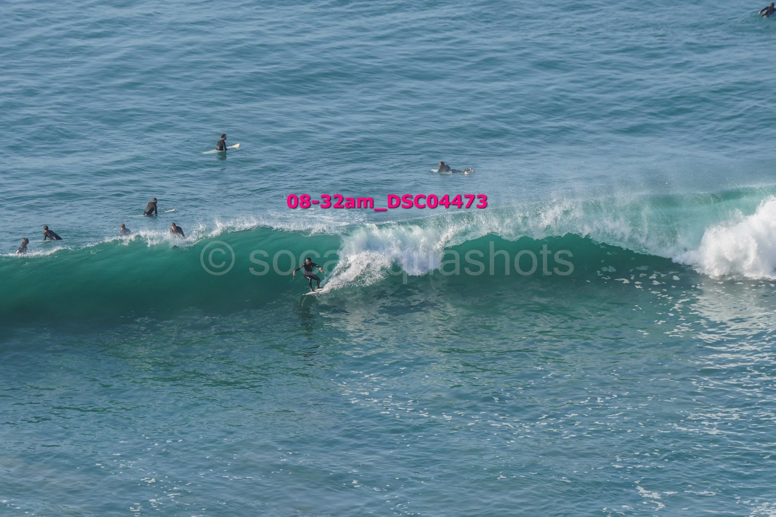 Surfer riding a wave with multiple surfers and swimmers in the water in the background.