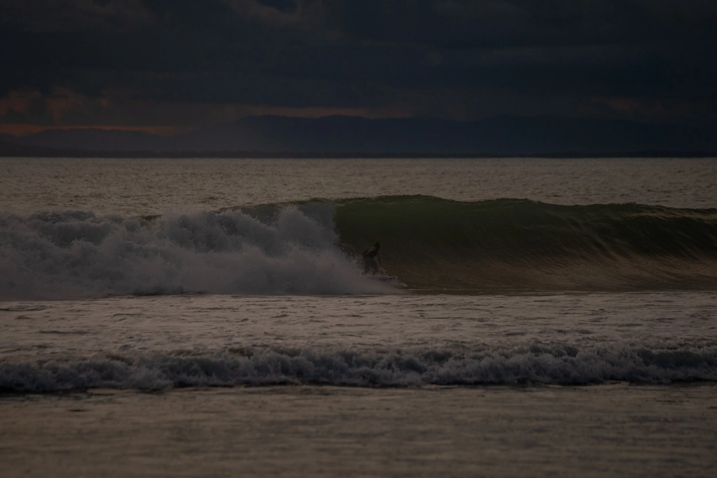 A person surfing on a wave during twilight, with a dark cloudy sky and mountains in the background.