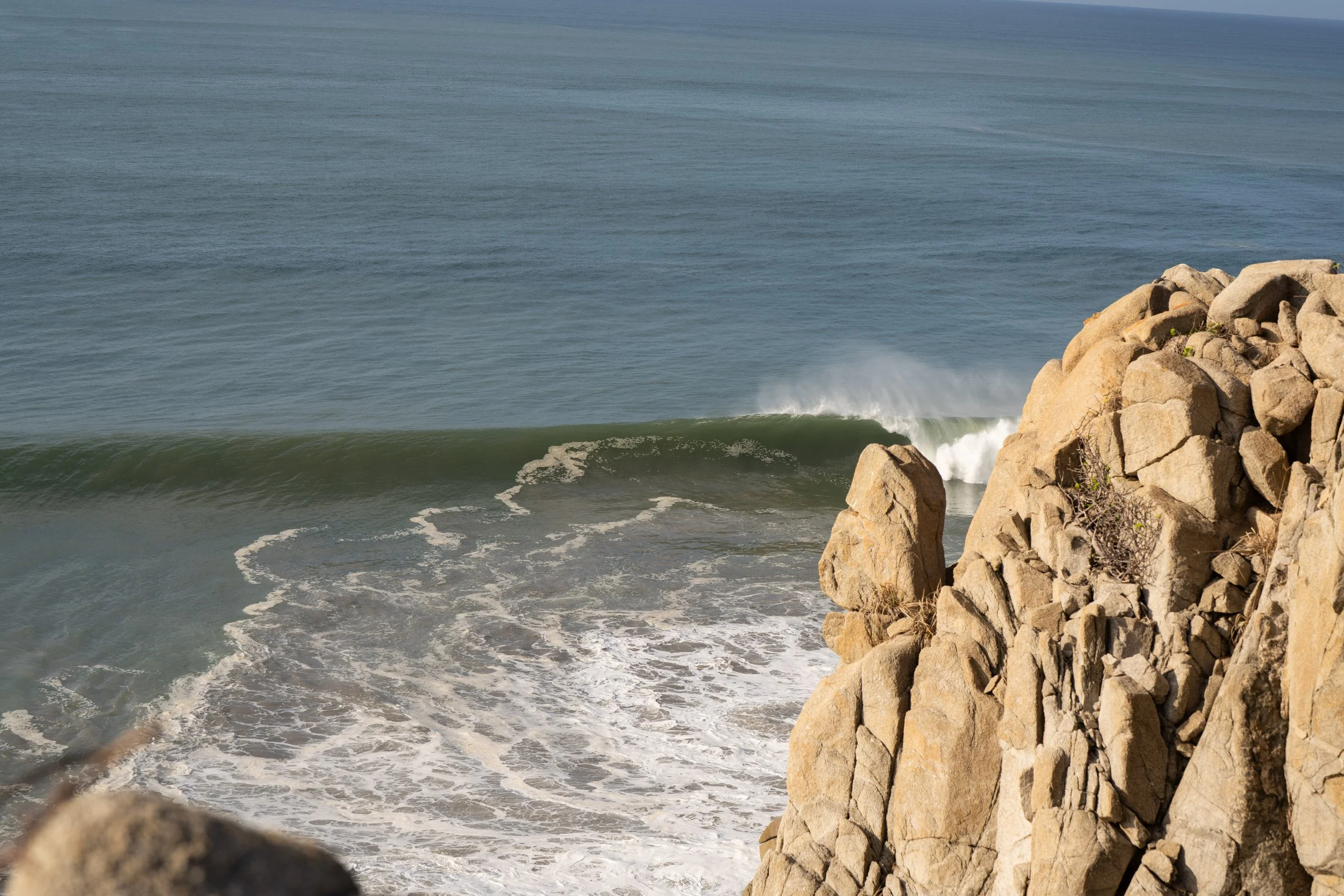 View of the ocean with waves breaking near large rocky cliffs on the right side, under a clear sky.
