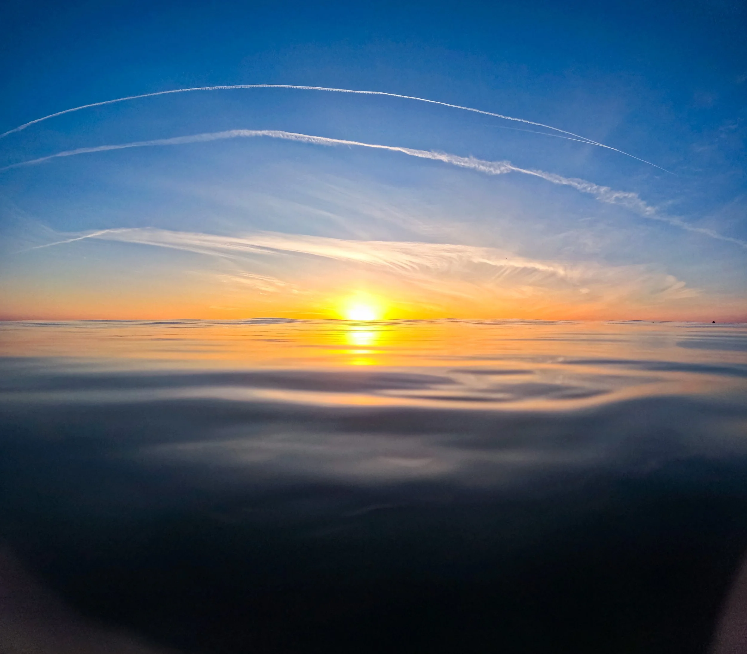 Sunset over calm ocean with colorful sky and jet trails.