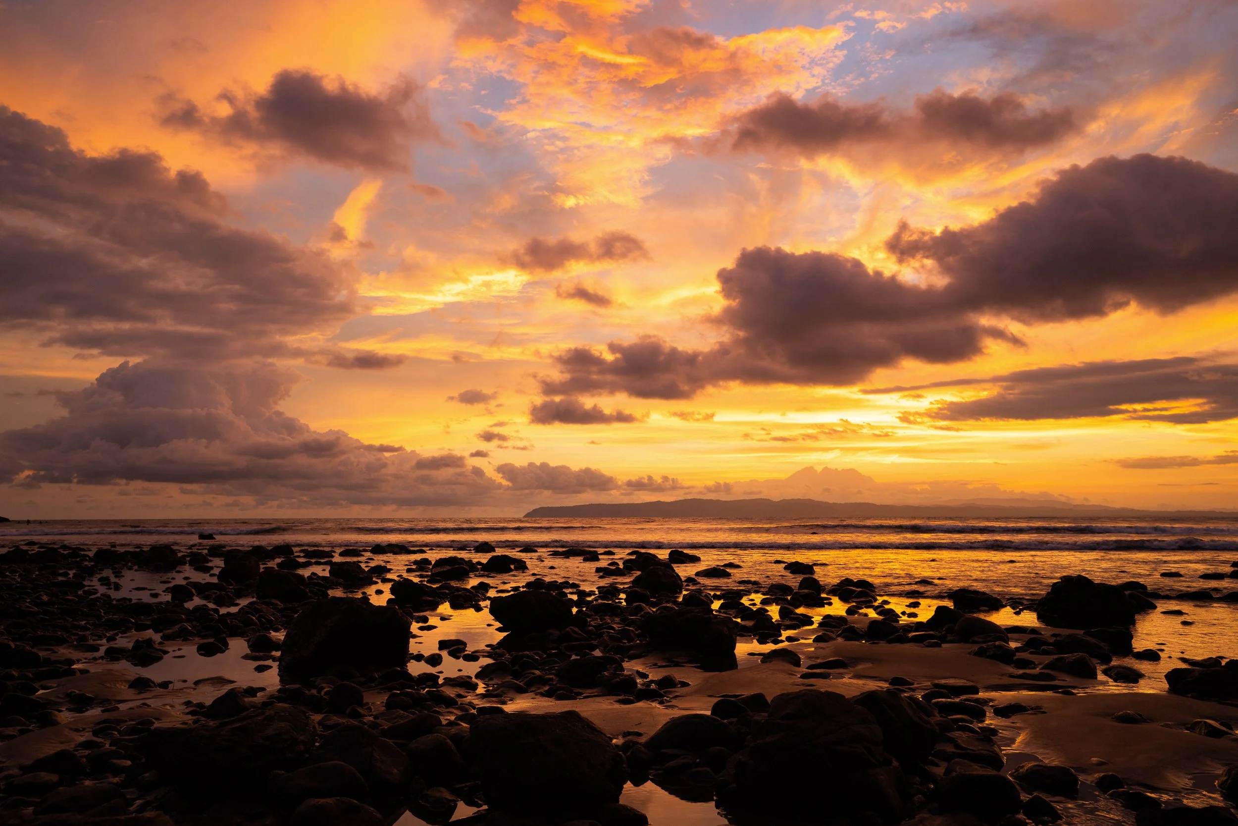 Sunset over the ocean with colorful clouds and a rocky beach in the foreground.