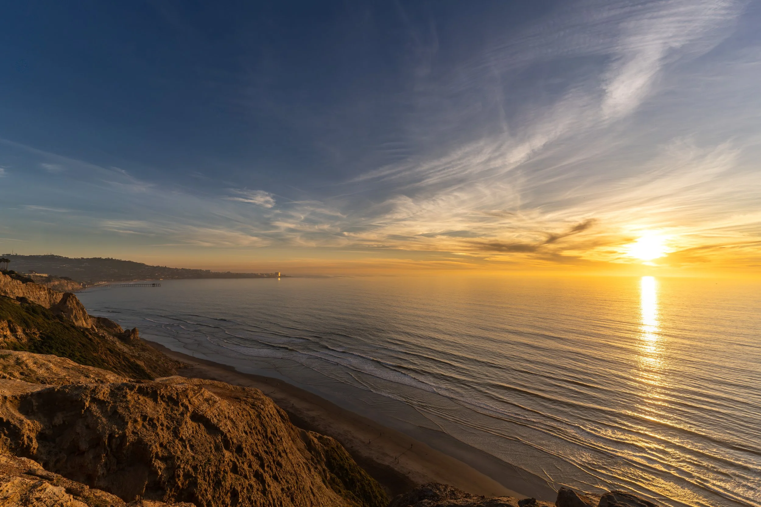 Sunset over the ocean with a sandy beach and rocky cliffs in the foreground.