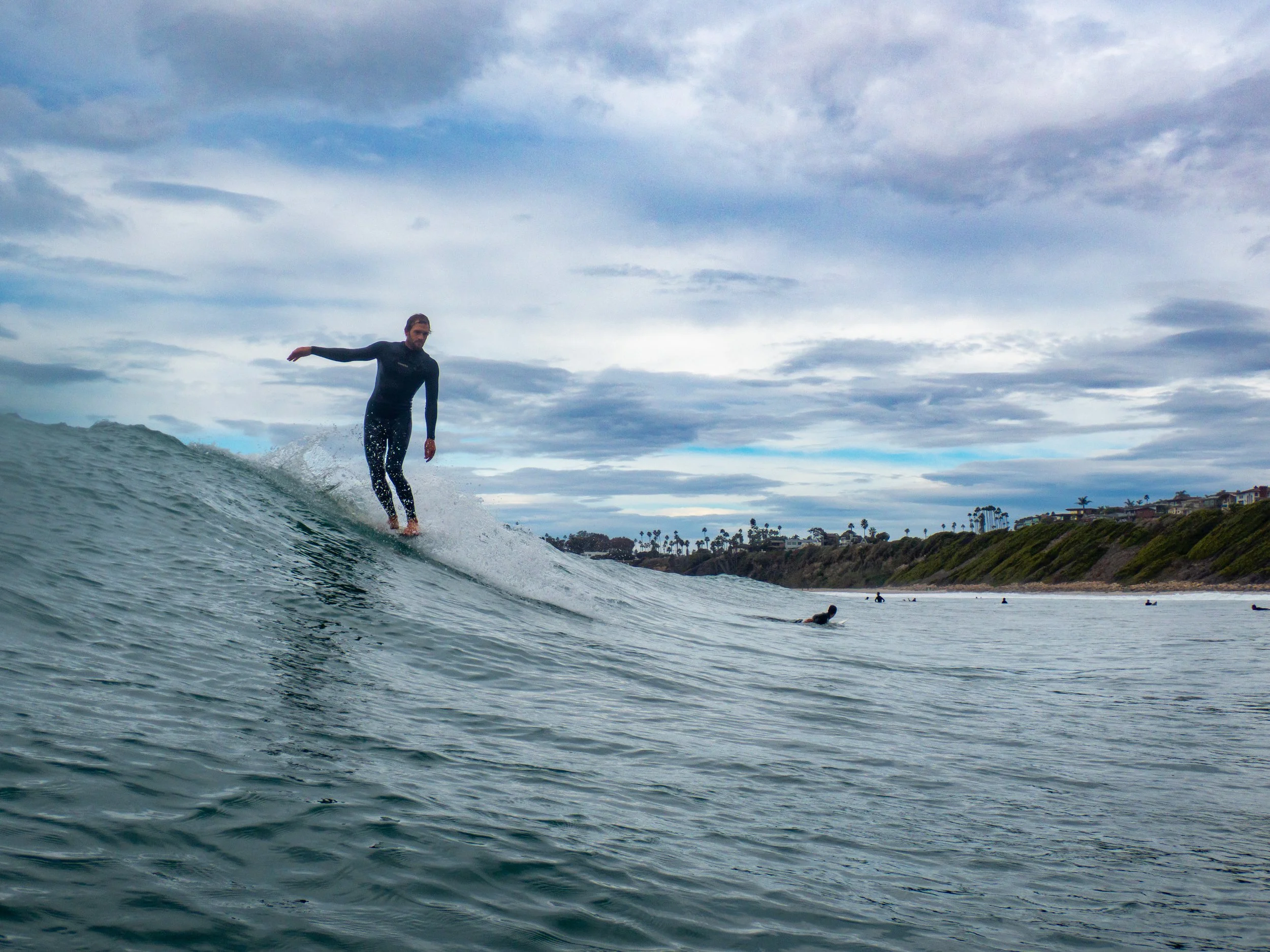 A person surfing on a wave in the ocean with a shoreline and houses in the background on a cloudy day.