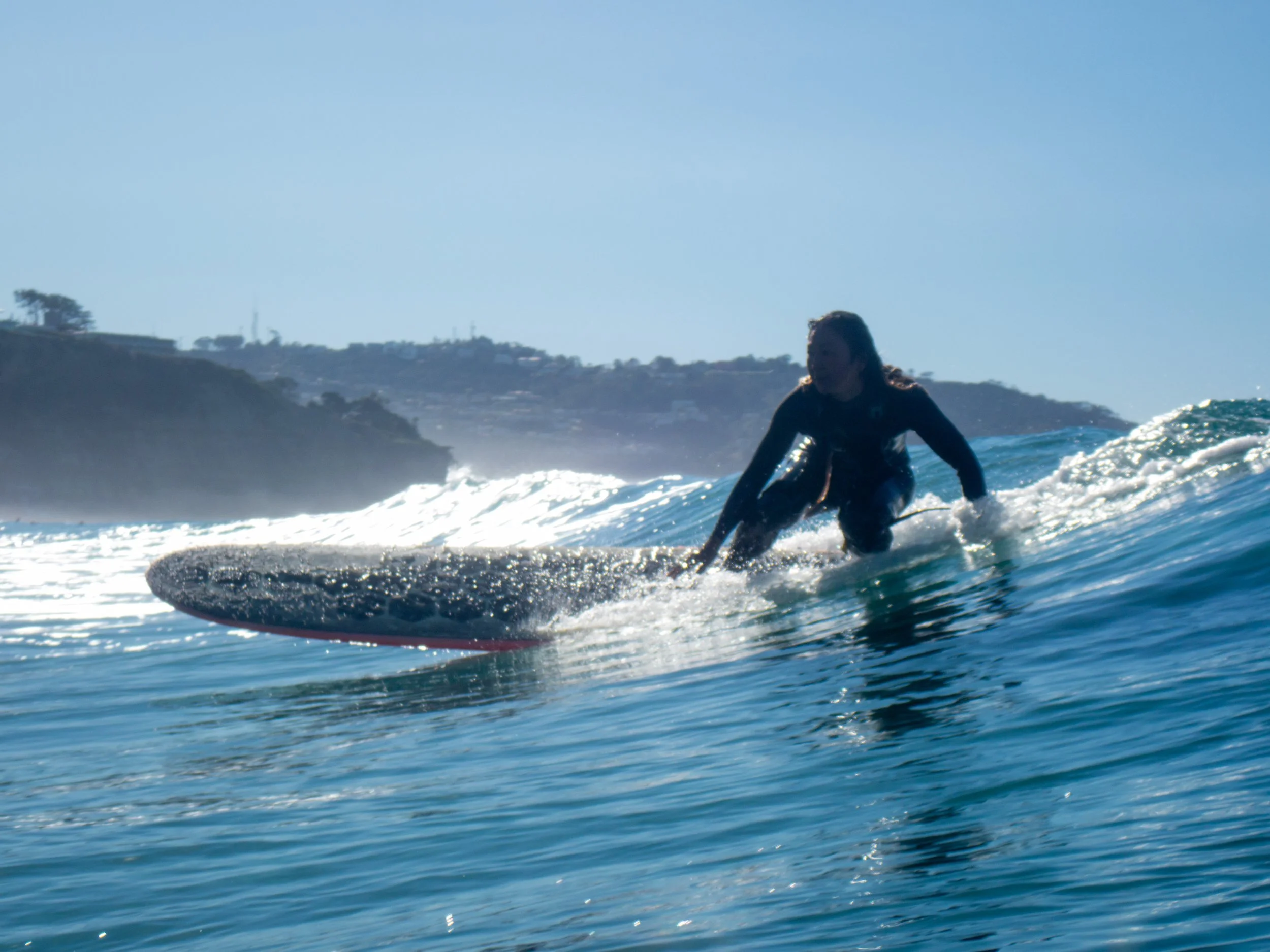 A person riding a wave on a surfboard in the ocean, with a coastline and hills in the background under a clear blue sky.