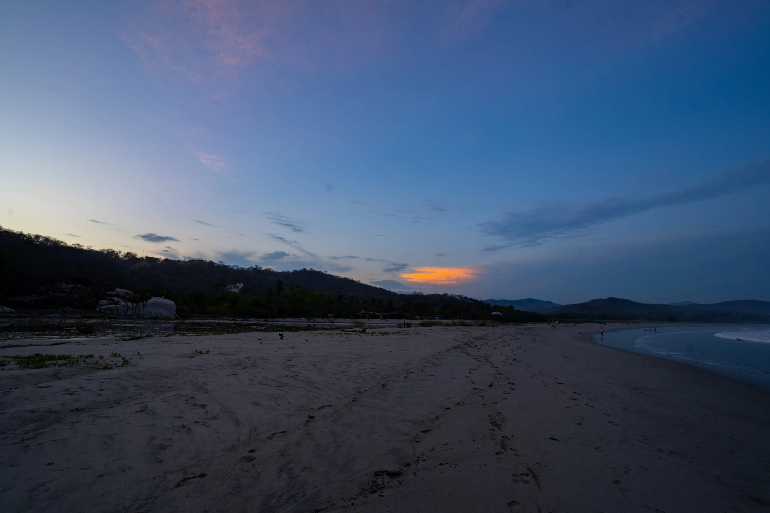 A beach during sunset with a calm sea, sand in the foreground, and hills covered with trees in the background. The sky is partly cloudy with shades of blue, purple, and orange.