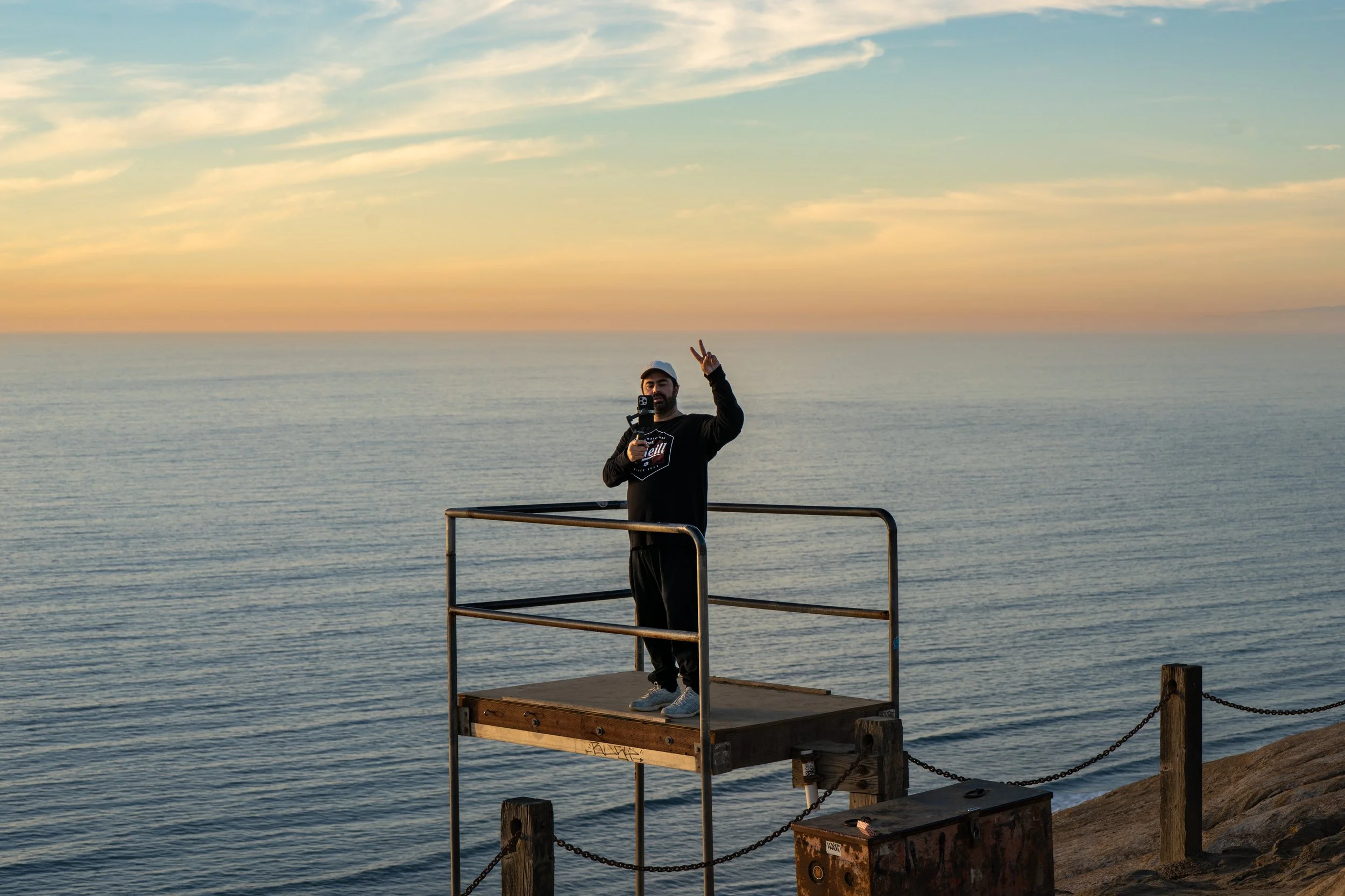 A man standing on a small elevated platform near the ocean holding a selfie stick and making a peace sign during sunset.