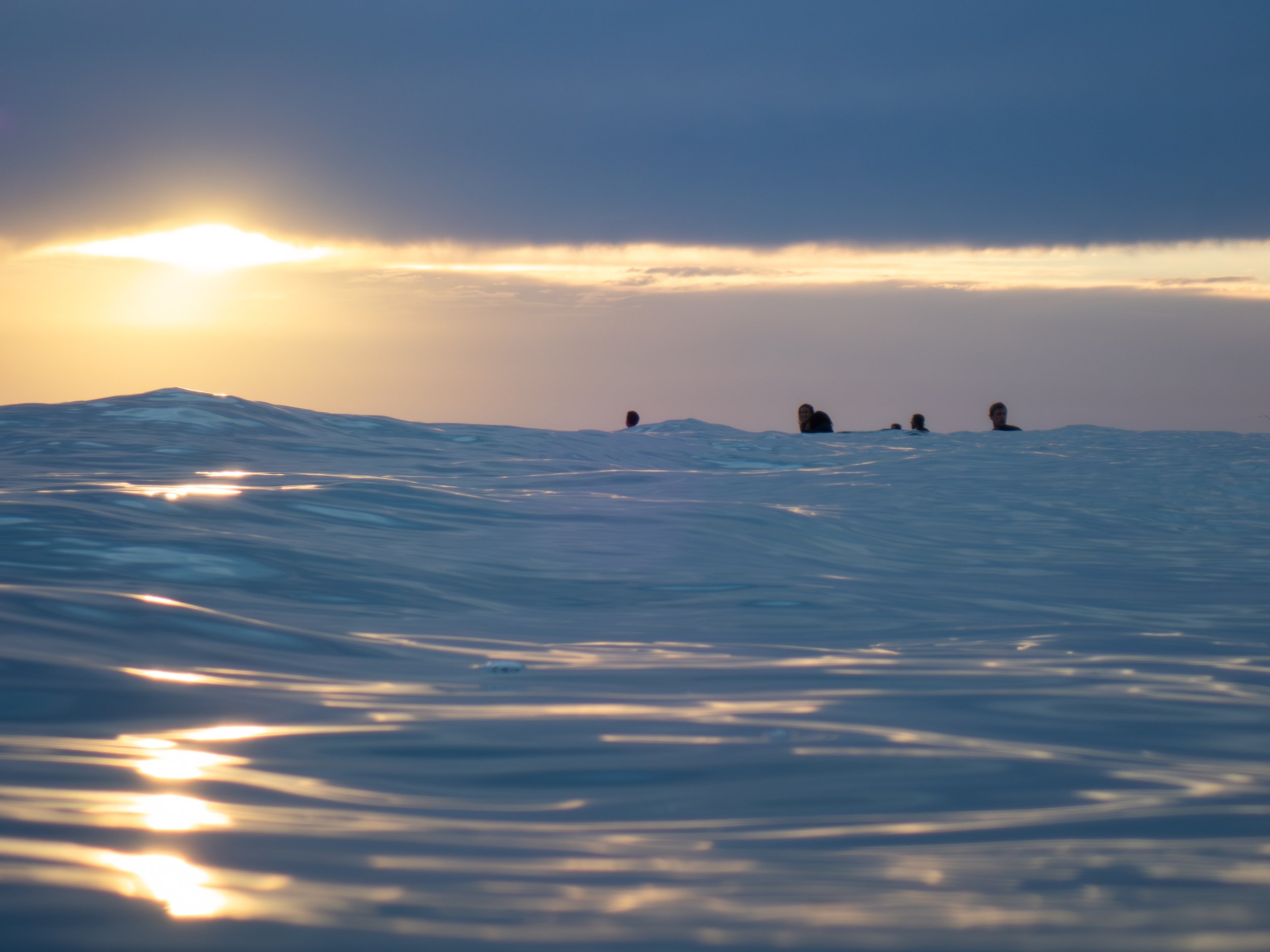 People in the ocean at sunset, with dark clouds overhead and sunlight reflecting on the water.