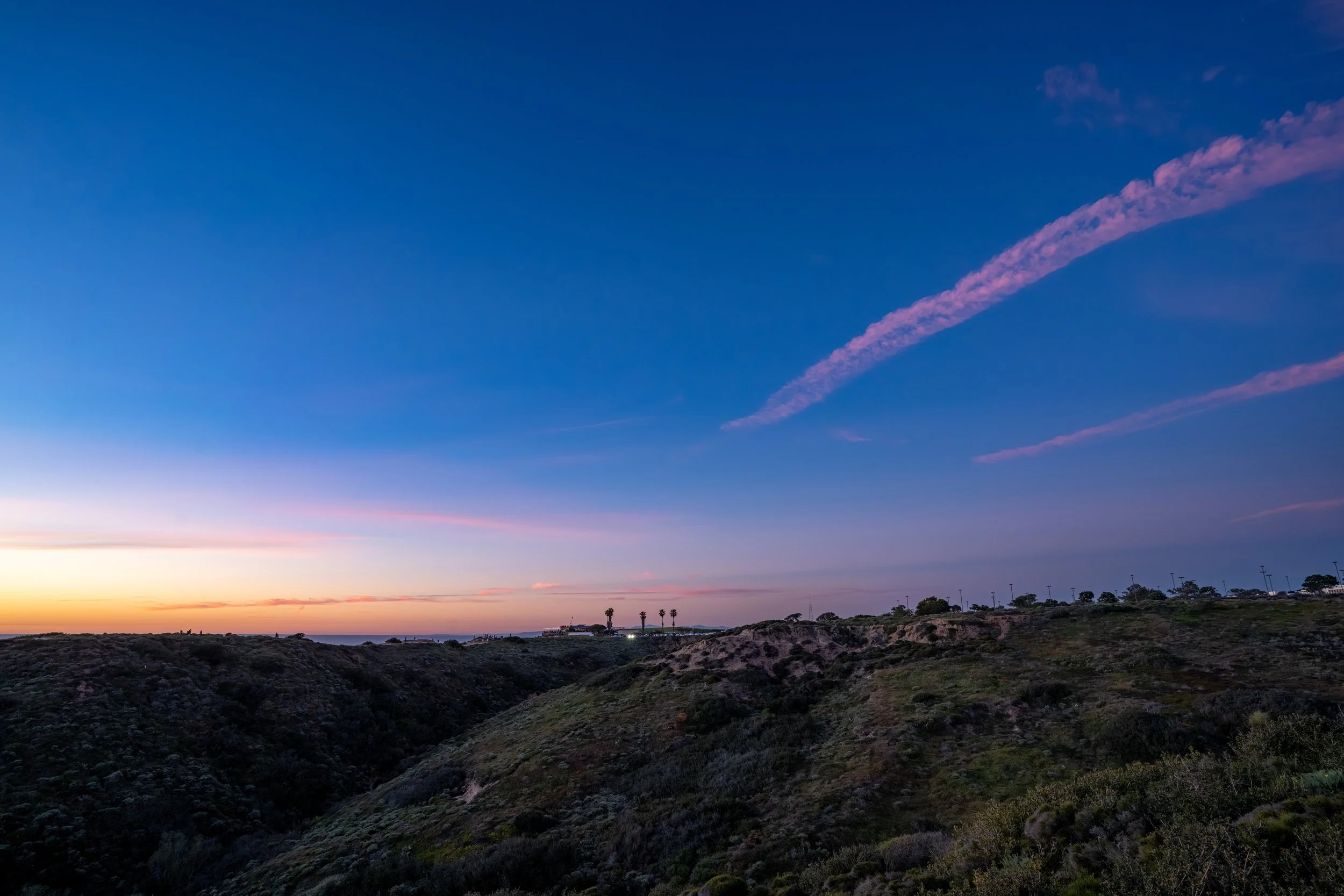 Sunset over a hilly landscape with sparse vegetation, with a few palm trees on the horizon and a mostly clear sky with a pink streak.