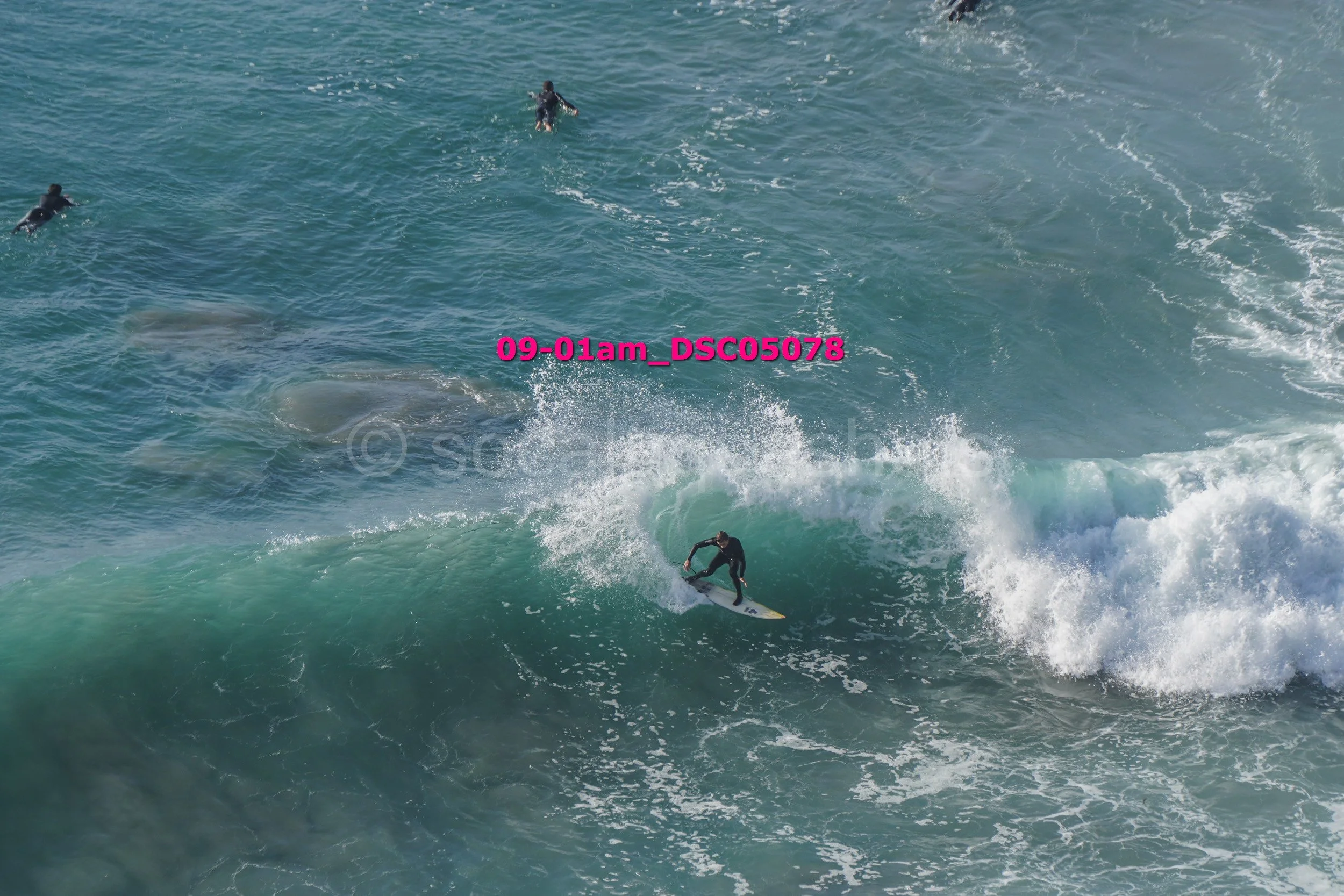 Surfer riding a wave with three other surfers in the water, seen from above.