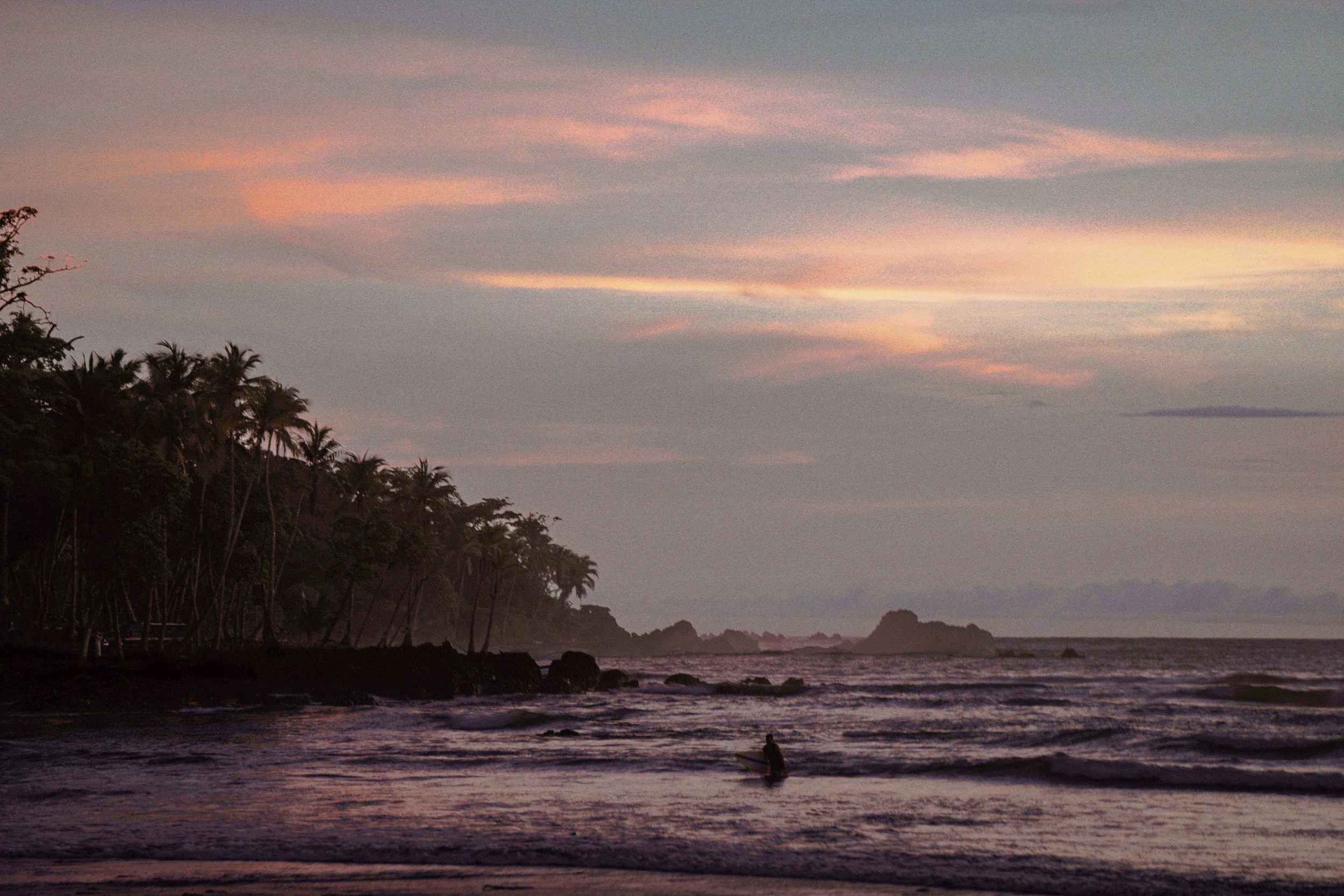 A sunset over a tropical beach with palm trees and a person paddleboarding on the water.