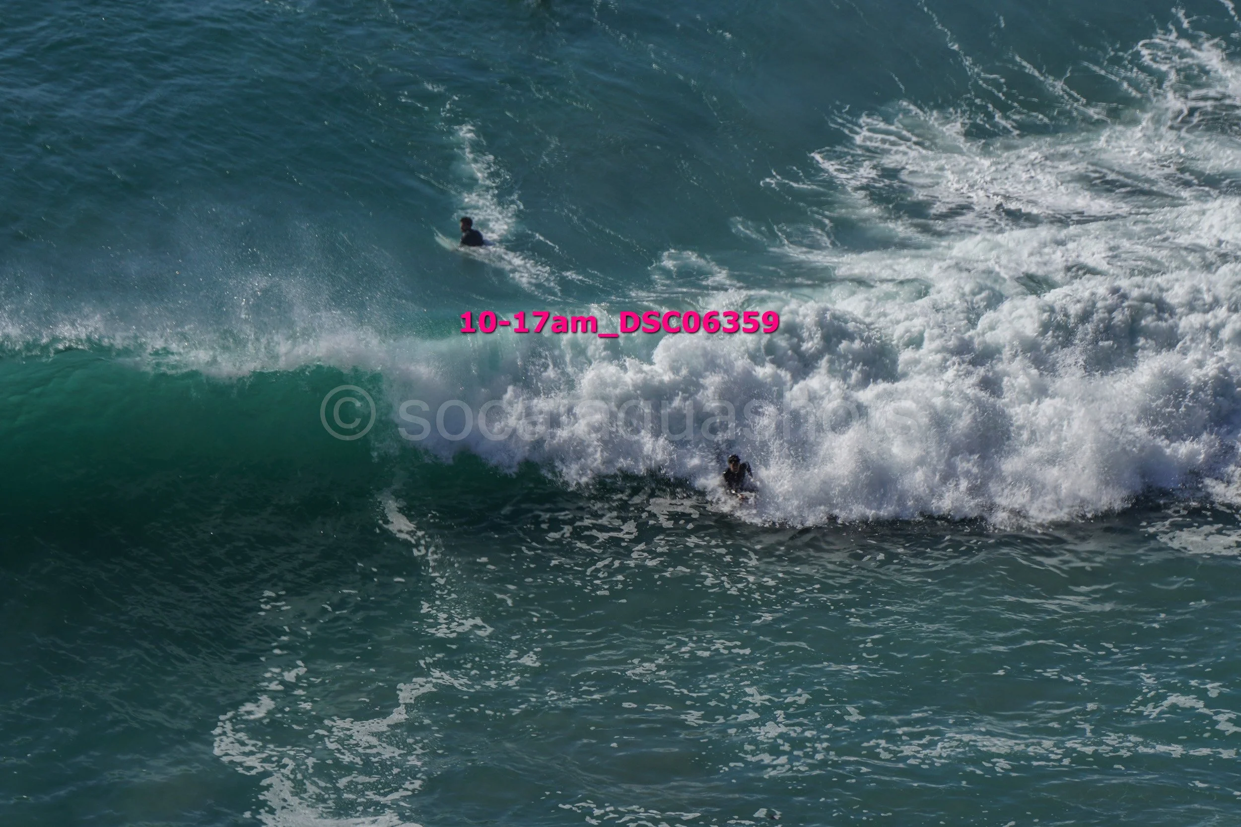 Two surfers riding large ocean waves, one partially submerged and one further back on the wave.