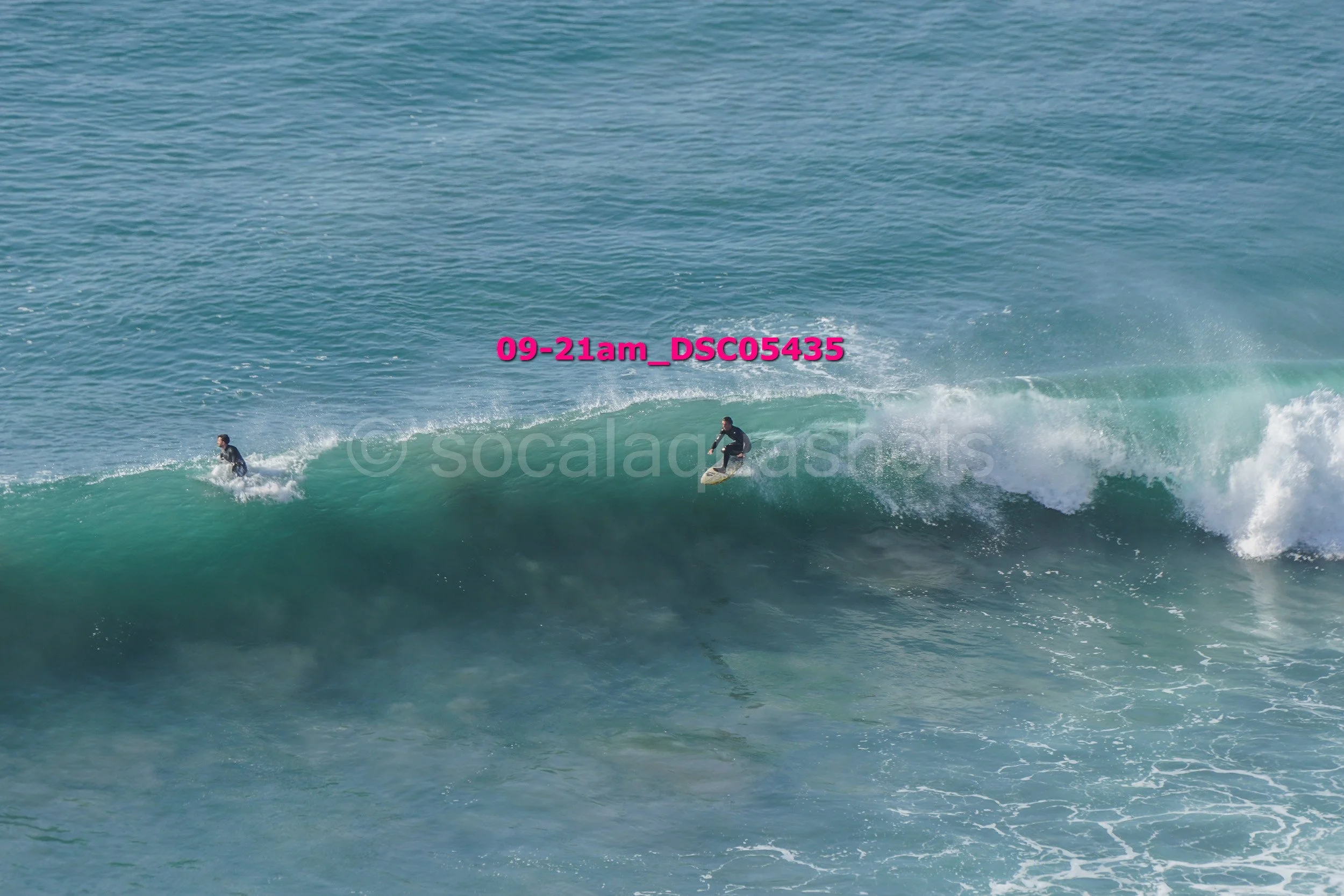 Two surfers riding a large wave in the ocean, with one surfer more towards the front and the other further back.