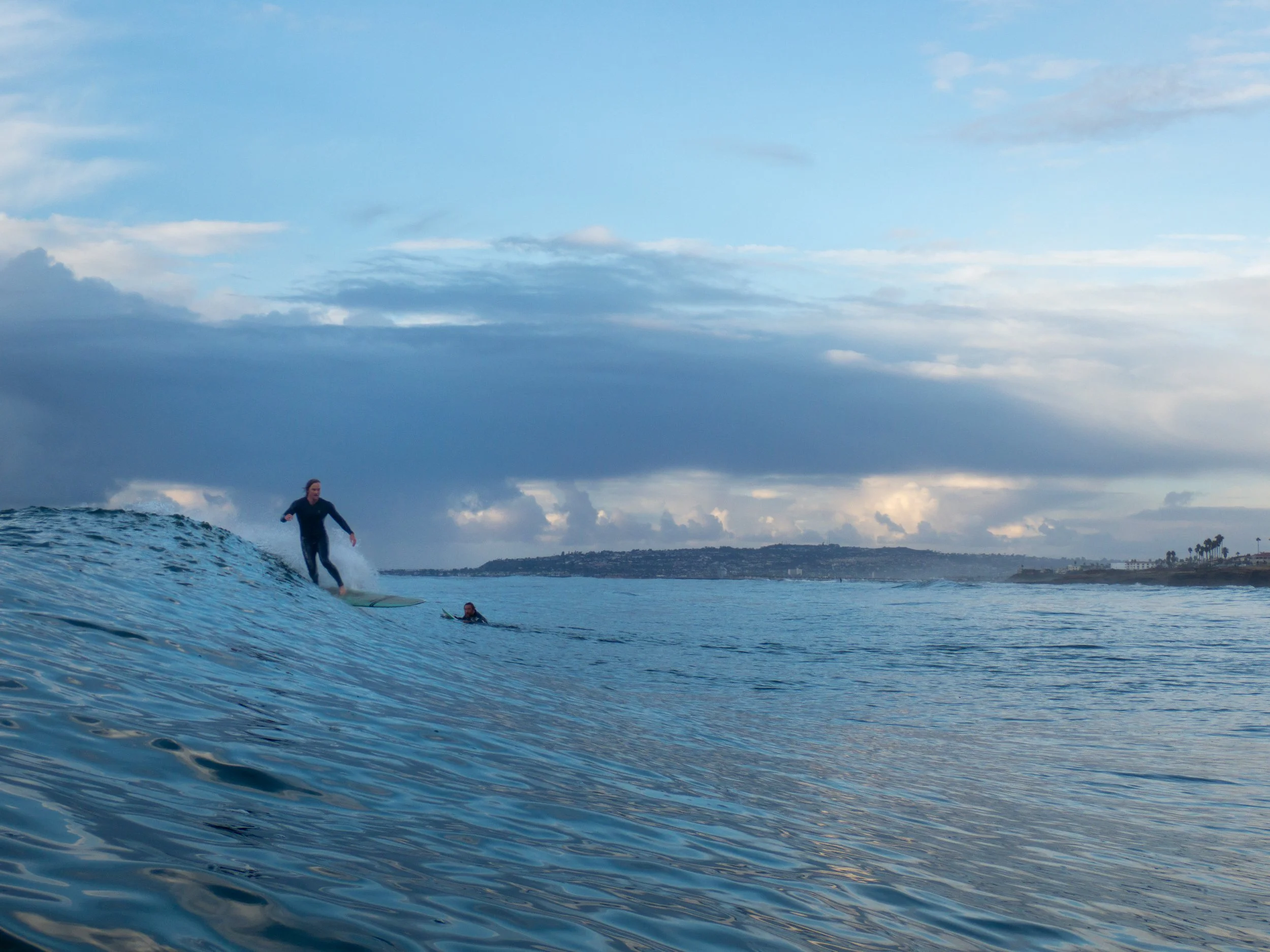 Person surfing on a wave near another person in the water with a coastline and cloudy sky in the background.
