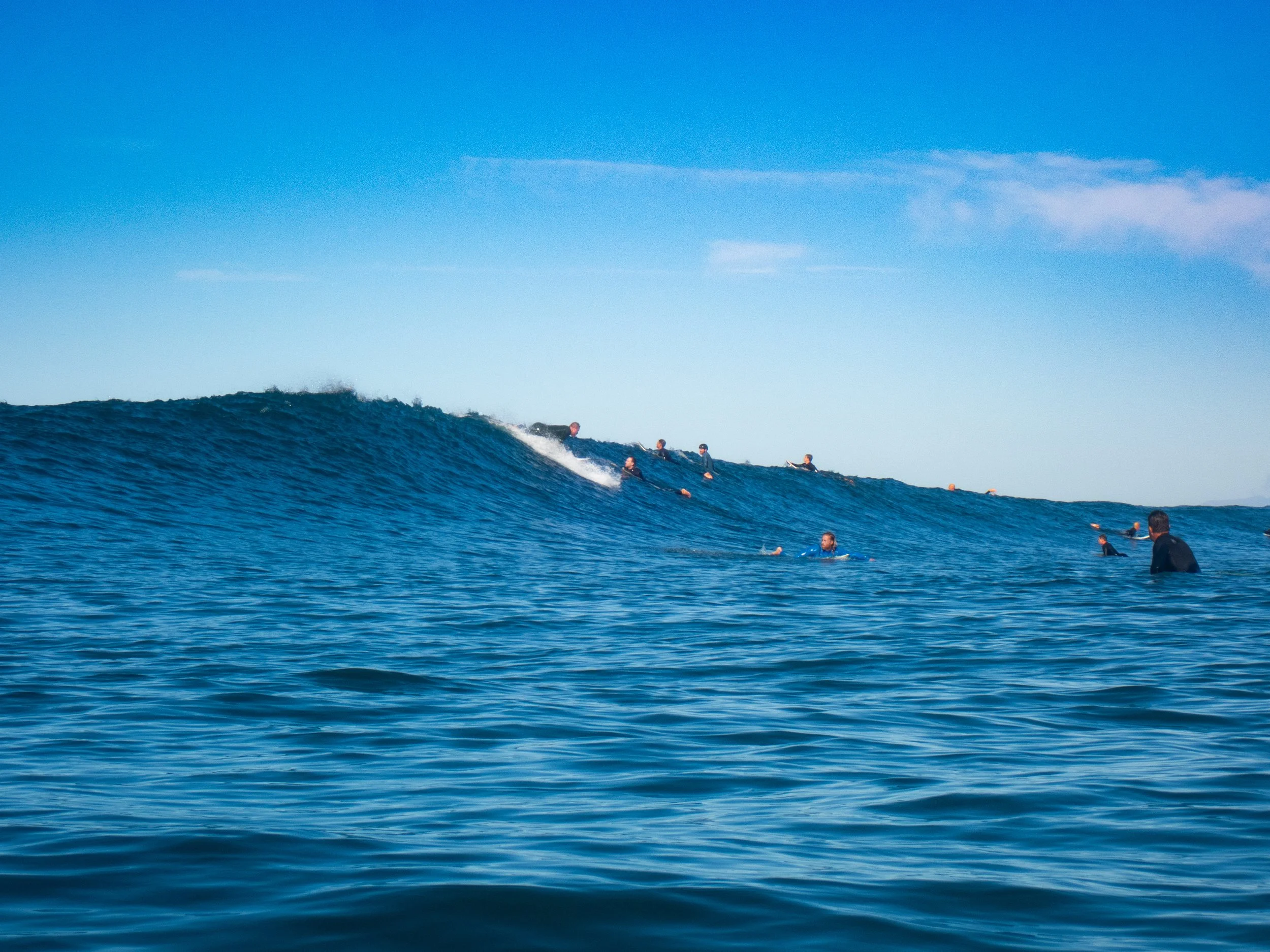 People surfing on a large blue wave in the ocean under a clear sky.
