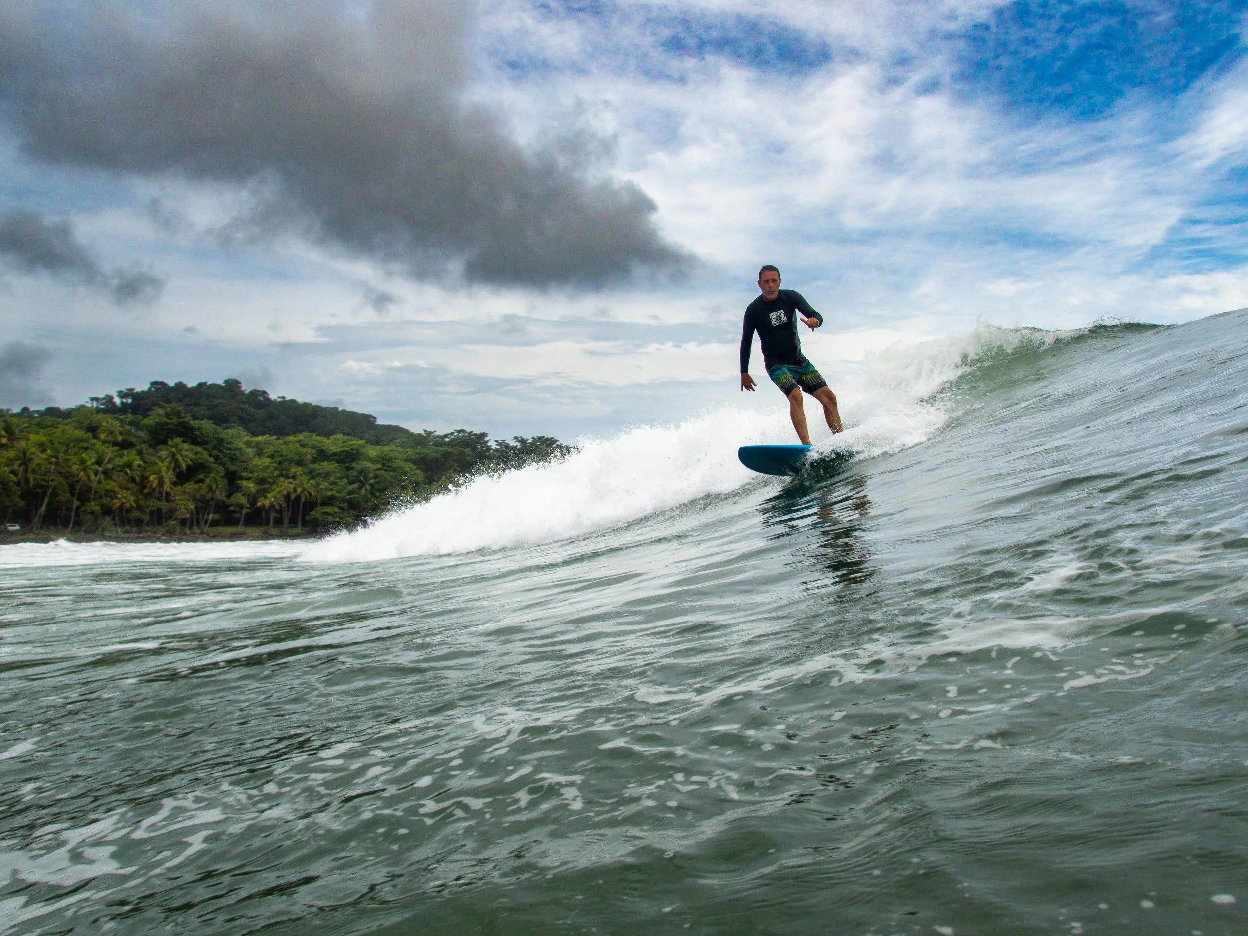 Surfer riding a wave with lush green forest and cloudy sky backdrop