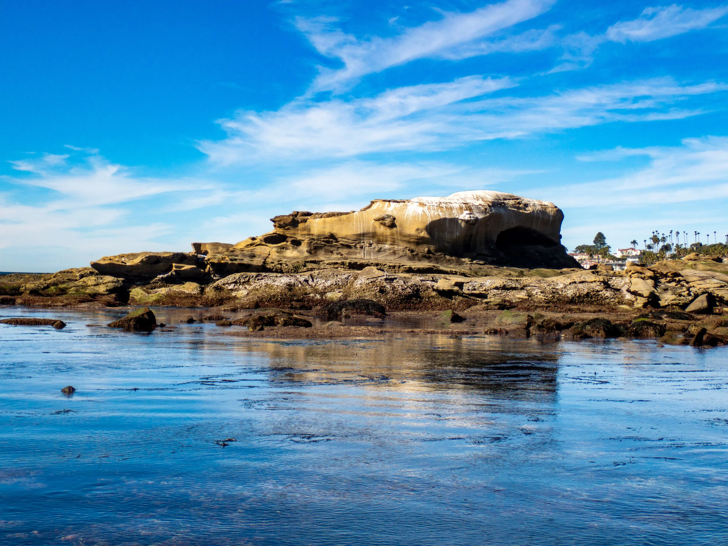 A large rock formation beside the ocean with a partly cloudy sky overhead and palm trees in the distance.