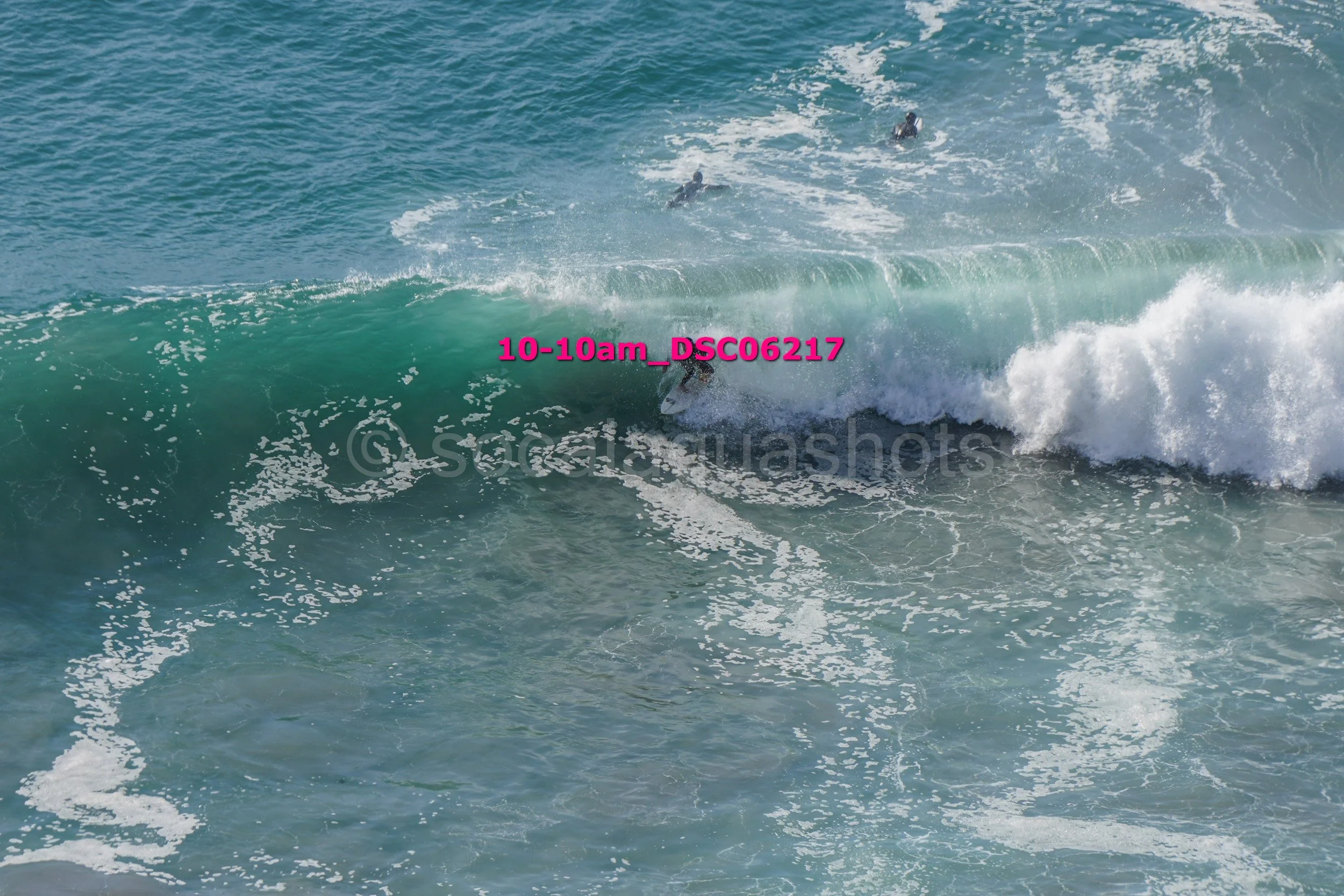 Surfer riding a green wave in the ocean with other surfers in the background.