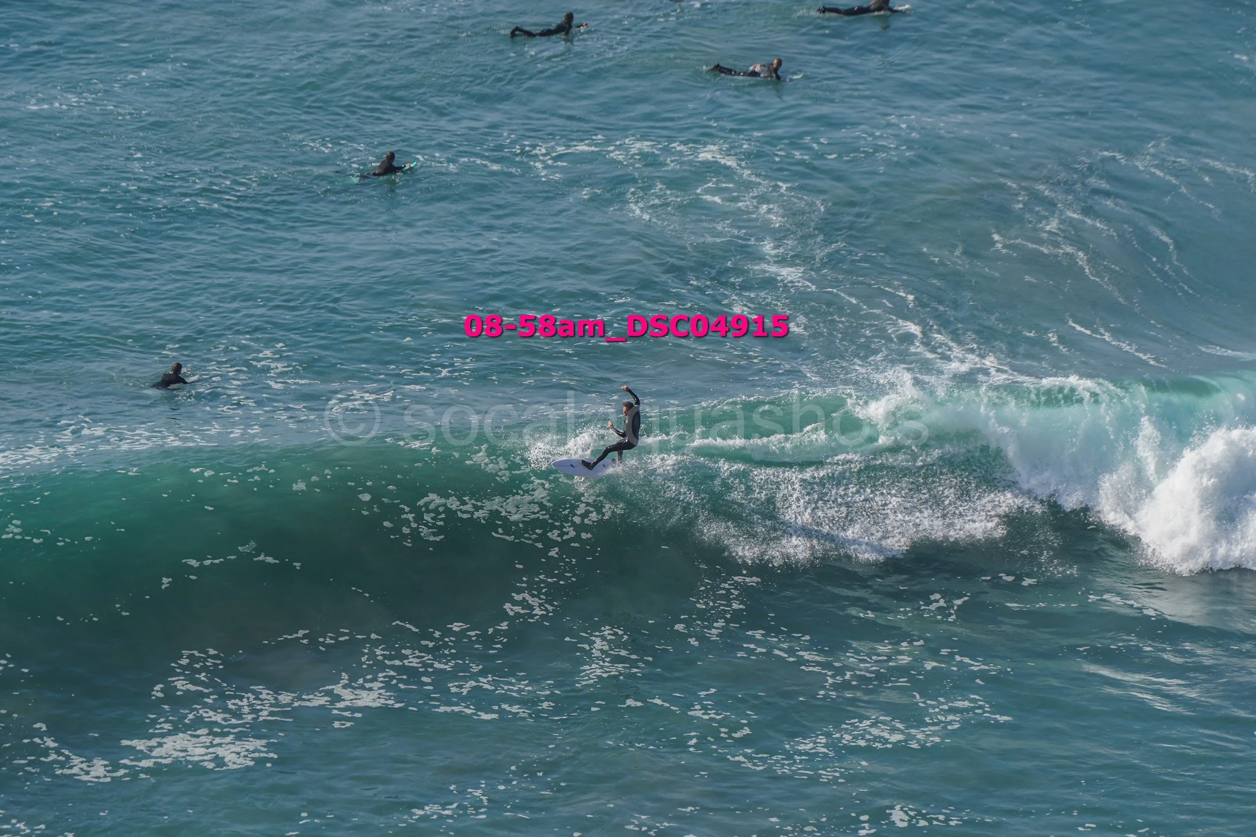 A surfer riding a wave in the ocean with several people swimming or surfing in the background.