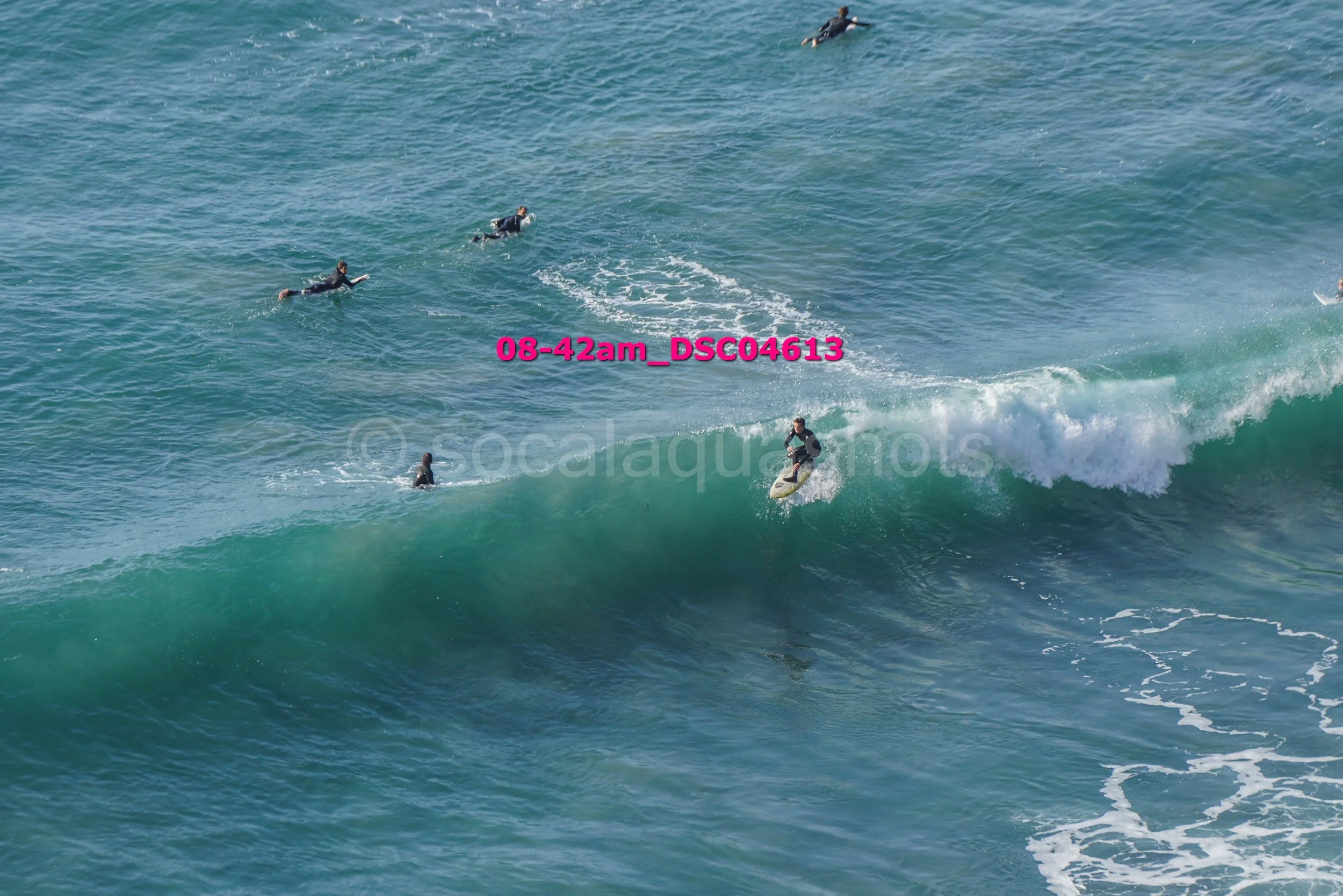Surfer riding a large ocean wave with other surfers swimming in the water nearby.