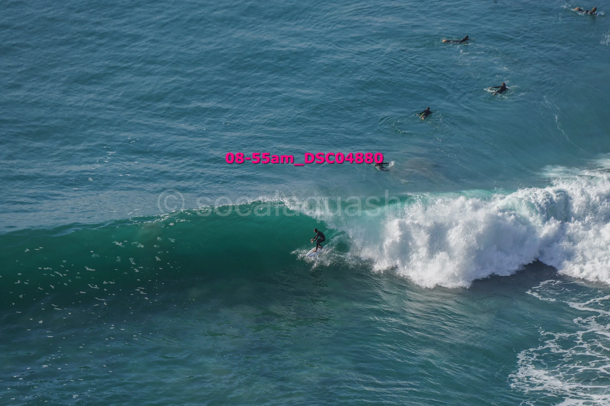 Surfer riding a wave in the ocean with several people swimming or surfing in the background.
