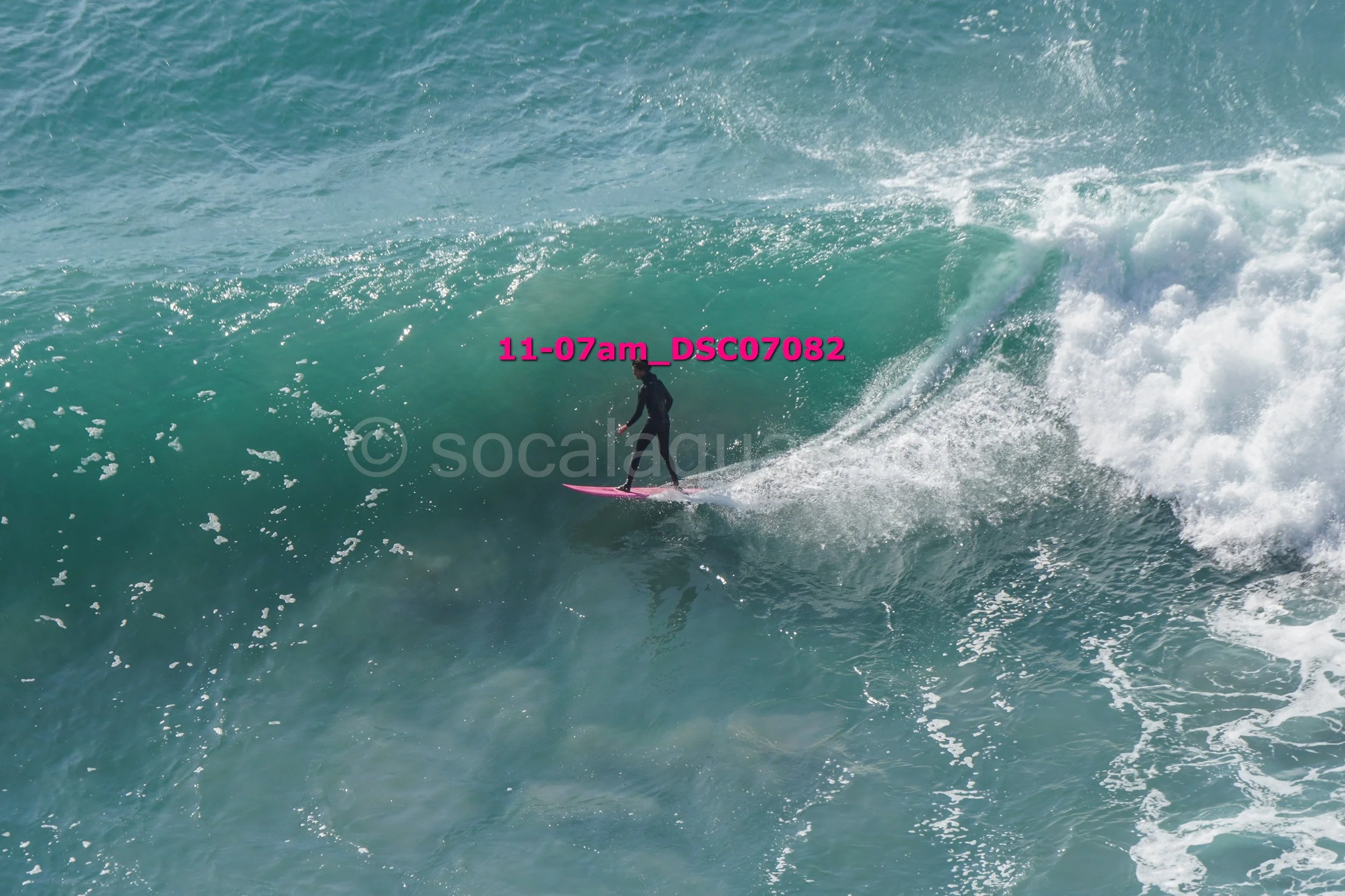 A person surfing on a pink surfboard on a large ocean wave with white foam, wearing a black wetsuit
