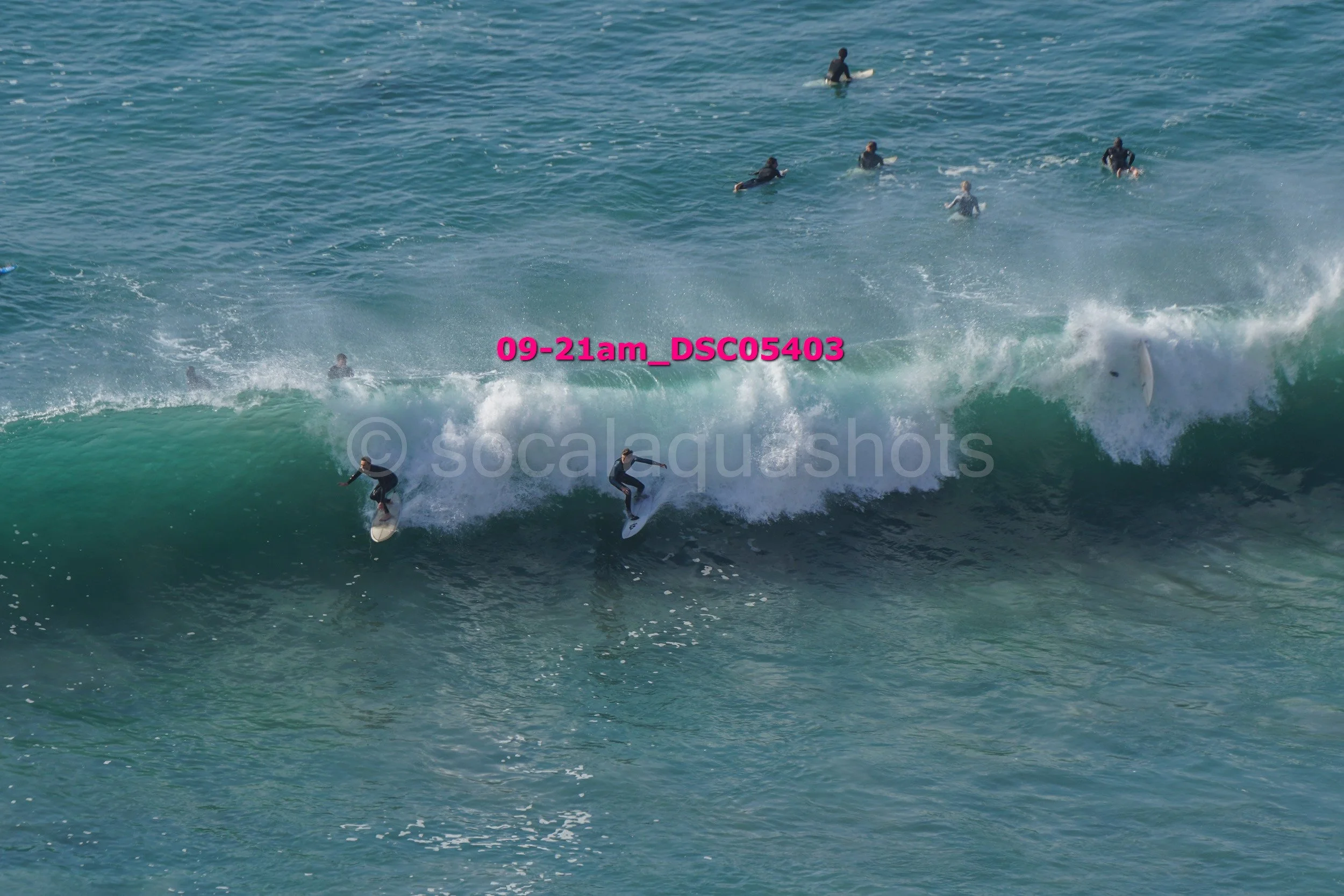 Surfers riding a large ocean wave with several more surfers in the background in the water.