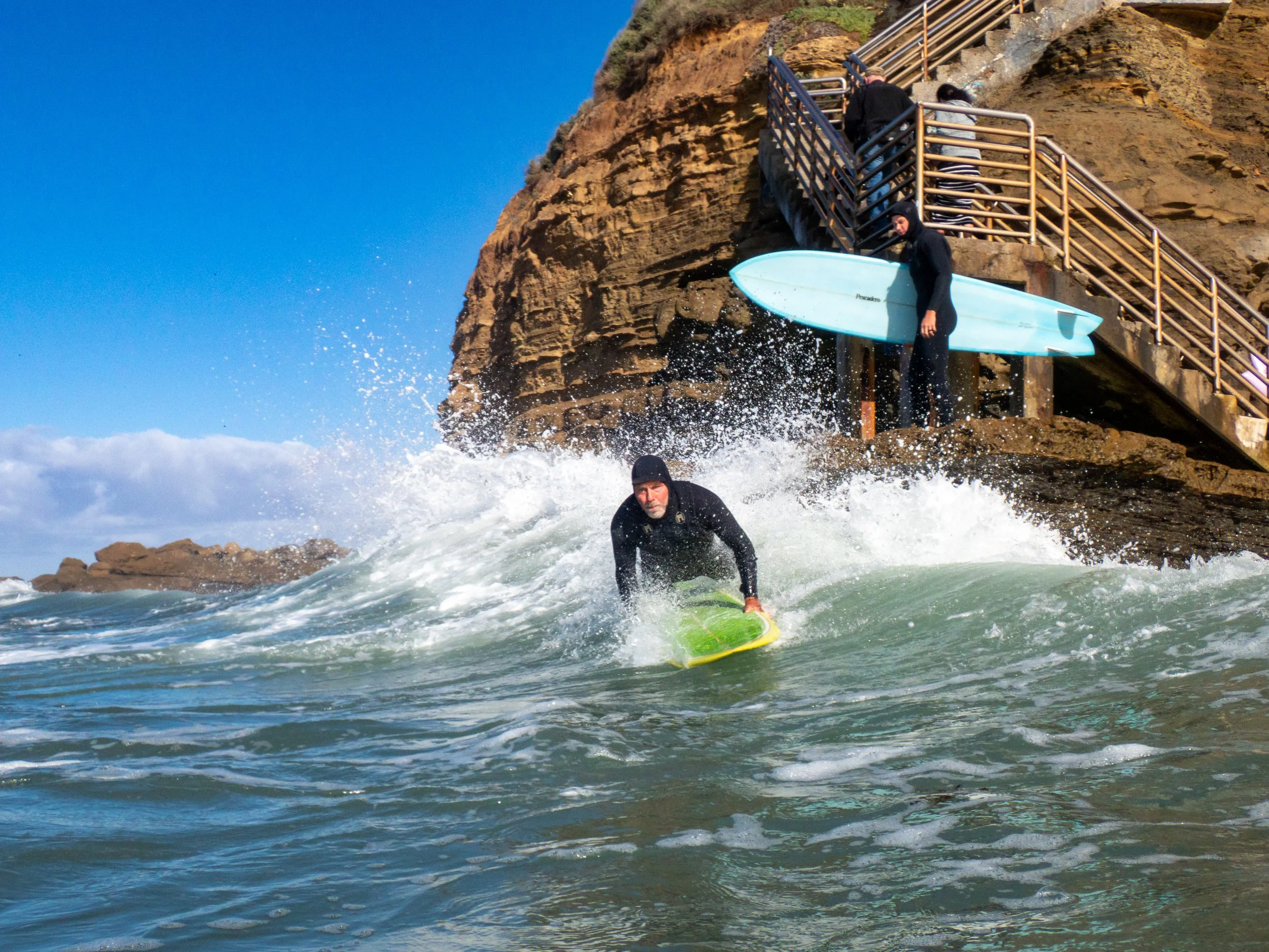 A person surfing on a green surfboard in the ocean near a rocky shore with a cliff and some people with surfboards and backpacks standing on a stairway on the cliff in the background.