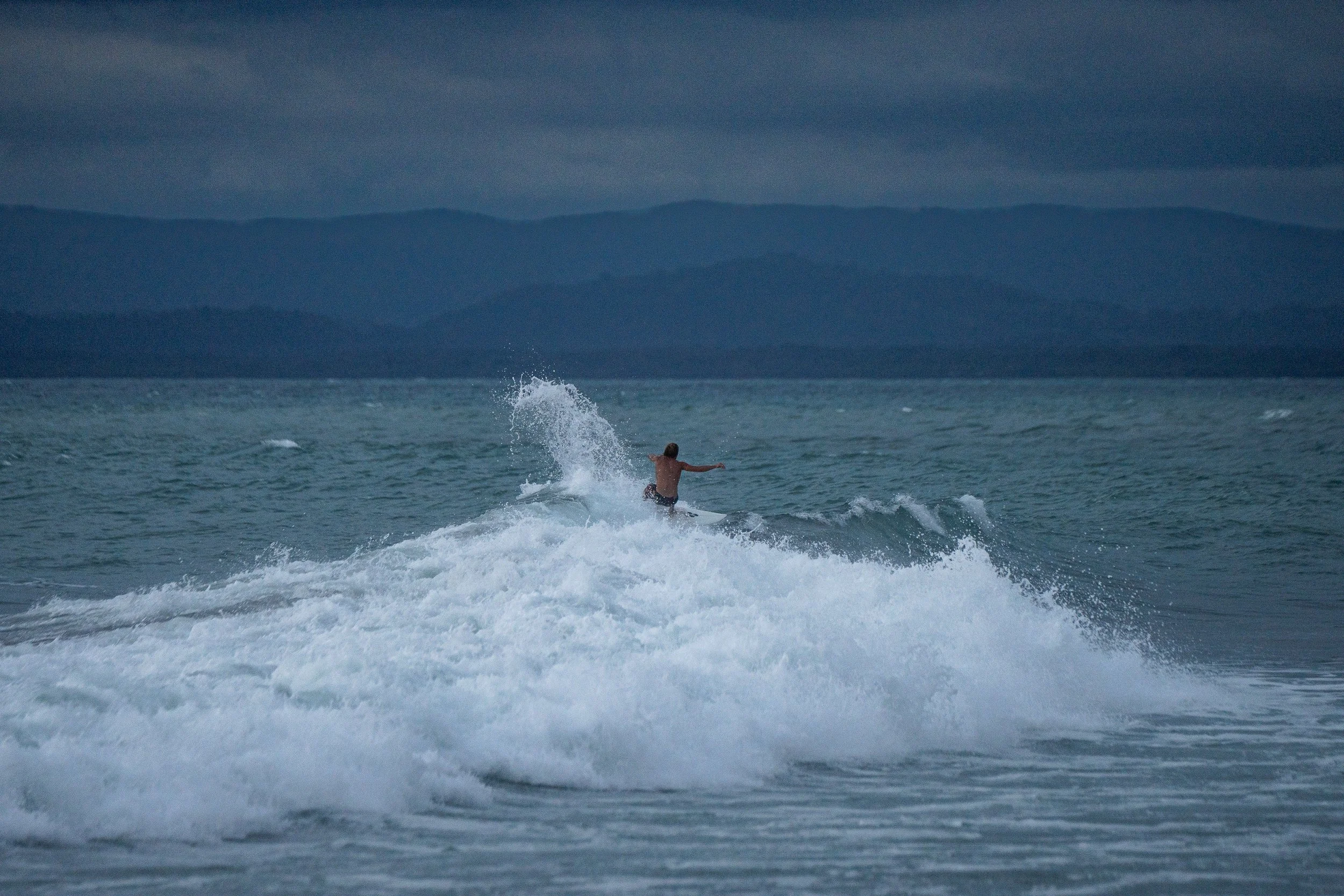 A person surfing on a wave in the ocean under cloudy skies with mountains in the background.
