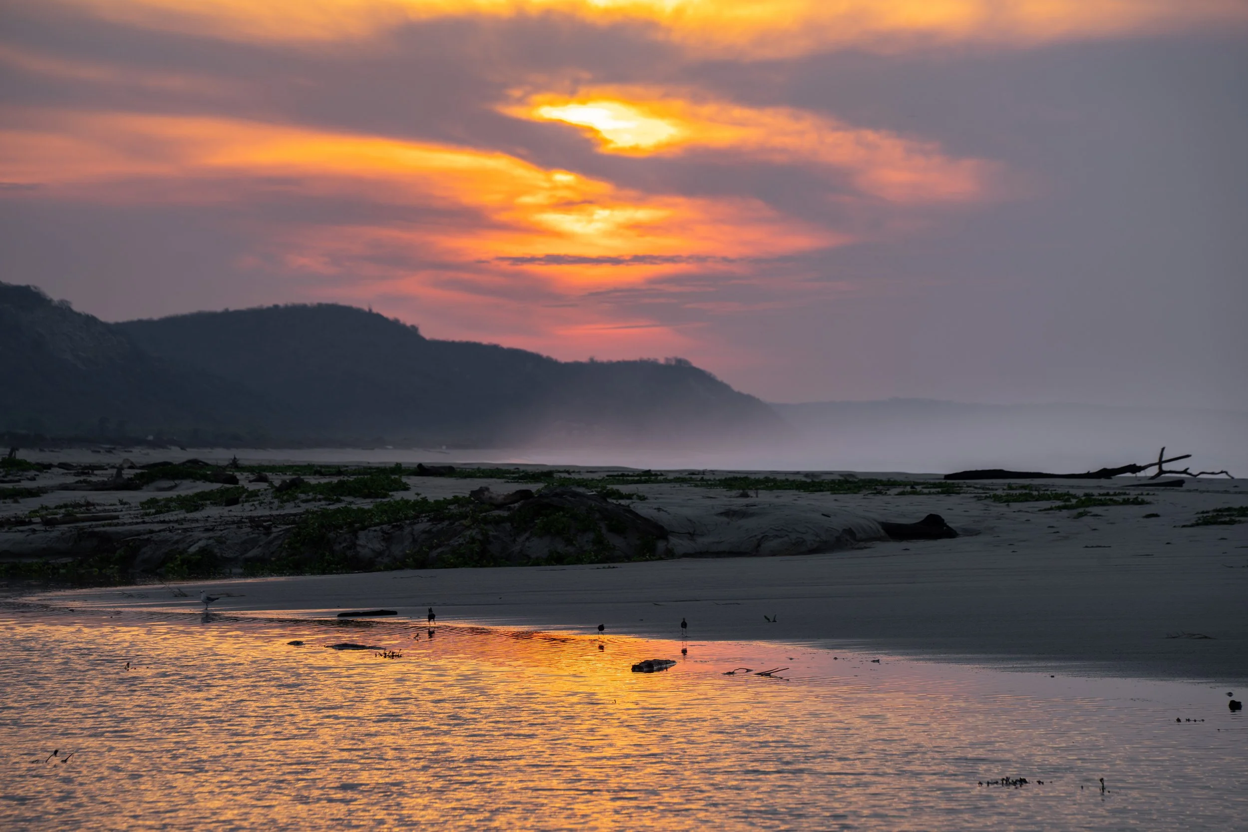 Sunset over a beach with mountains in the background, reflecting orange, pink, and yellow hues on the water.