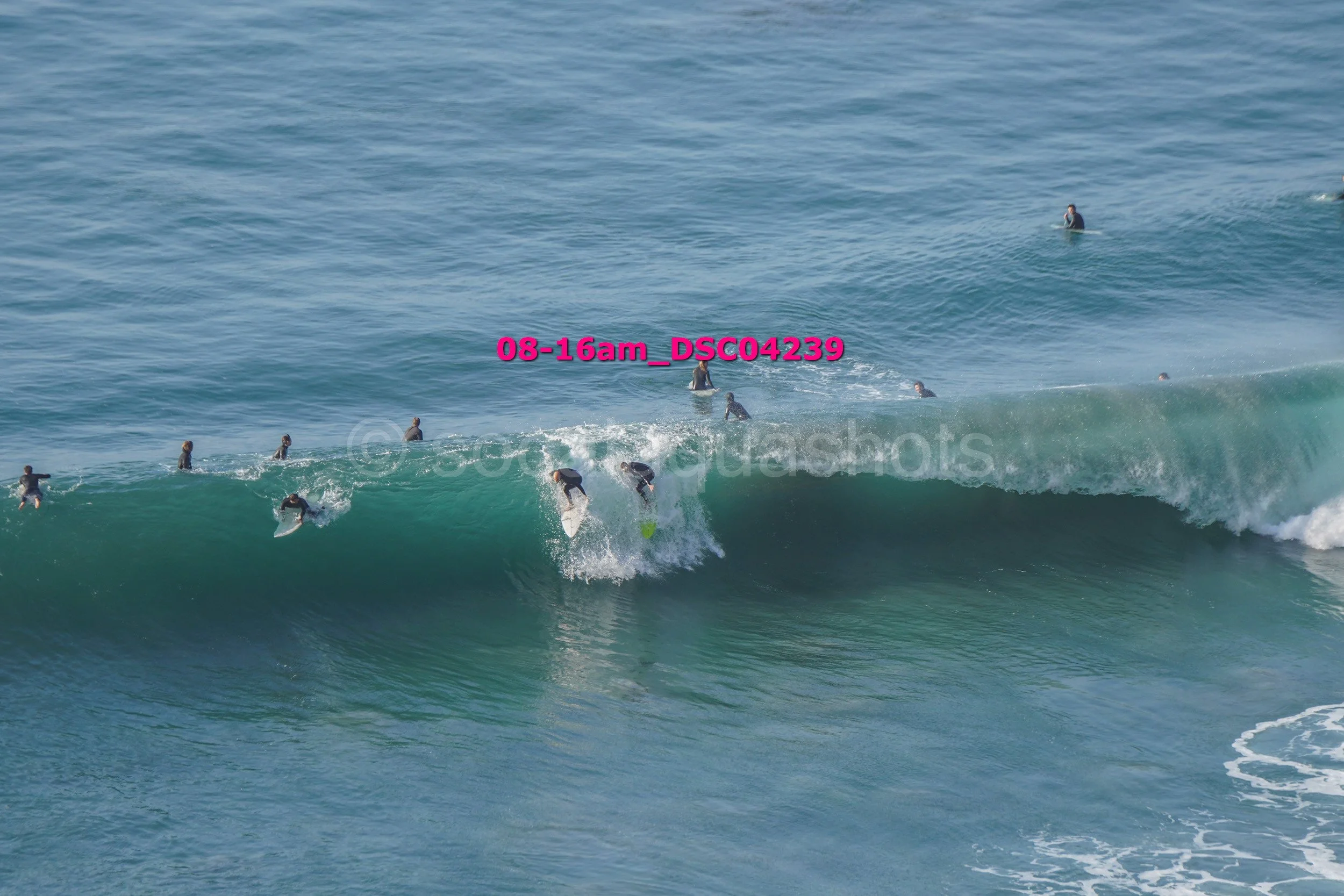 Multiple surfers in wetsuits riding and waiting for waves in the ocean, with clear blue water and a large breaking wave.