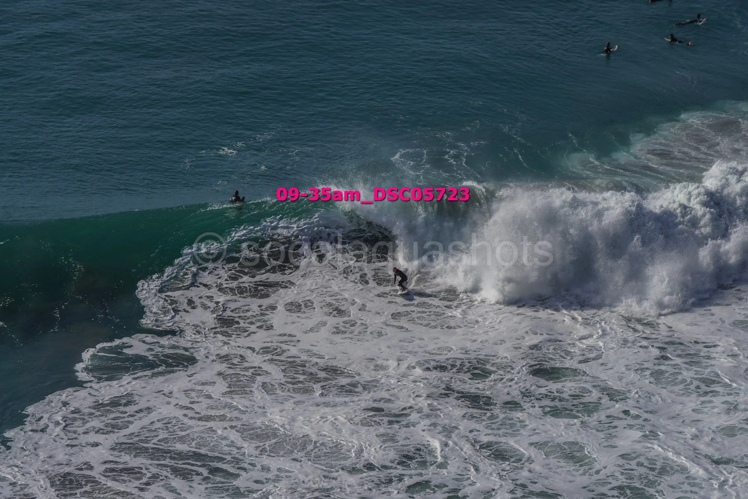Surfer riding a large wave with several other surfers in the water behind.