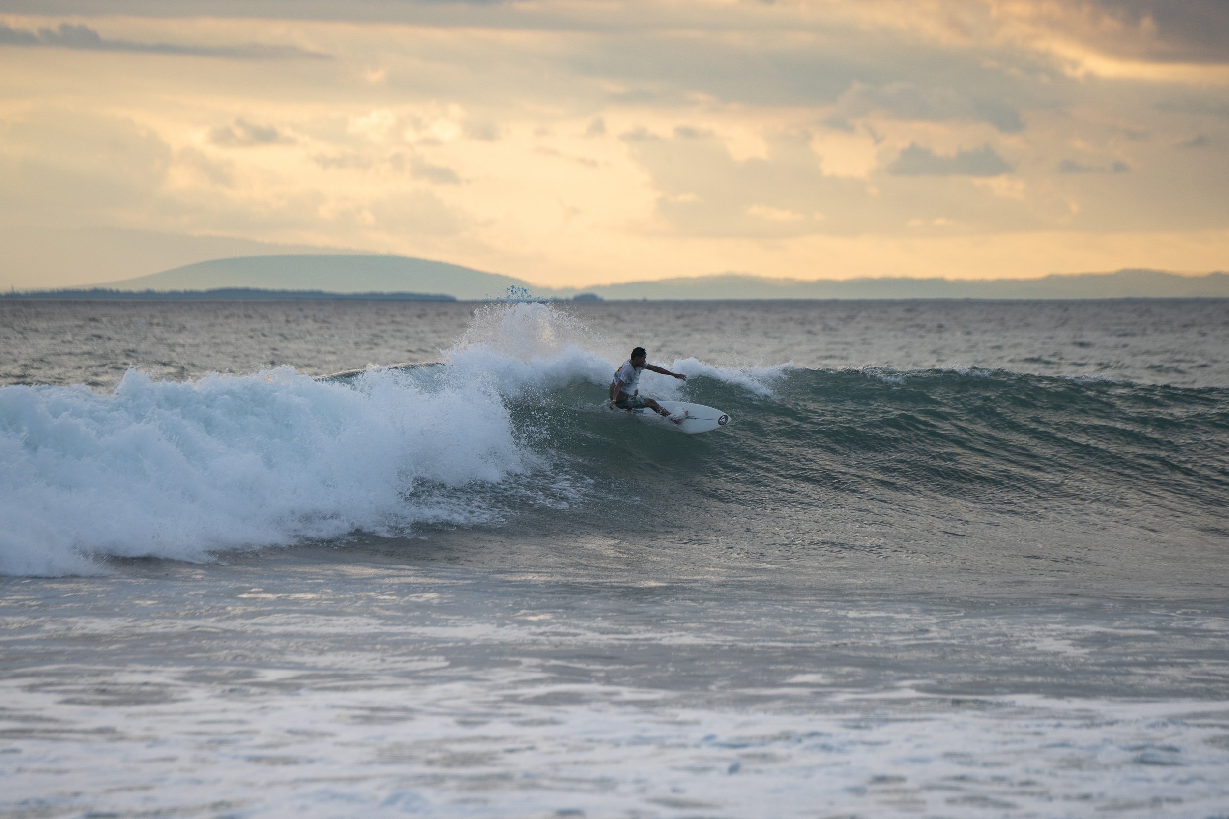 A person surfing on a wave in the ocean during sunset or sunrise with a cloudy sky and distant land in the background.
