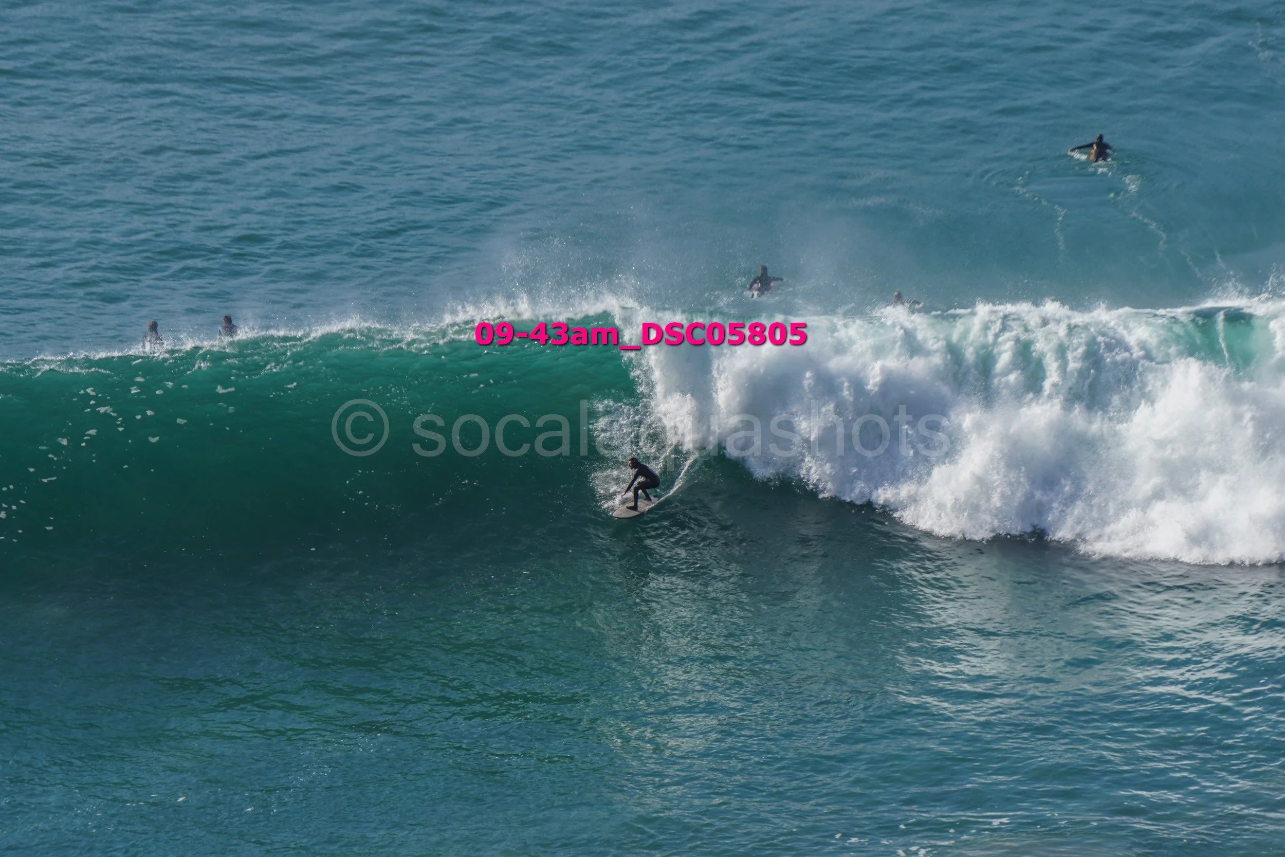 Surfer riding a large ocean wave while several people in the water watch.