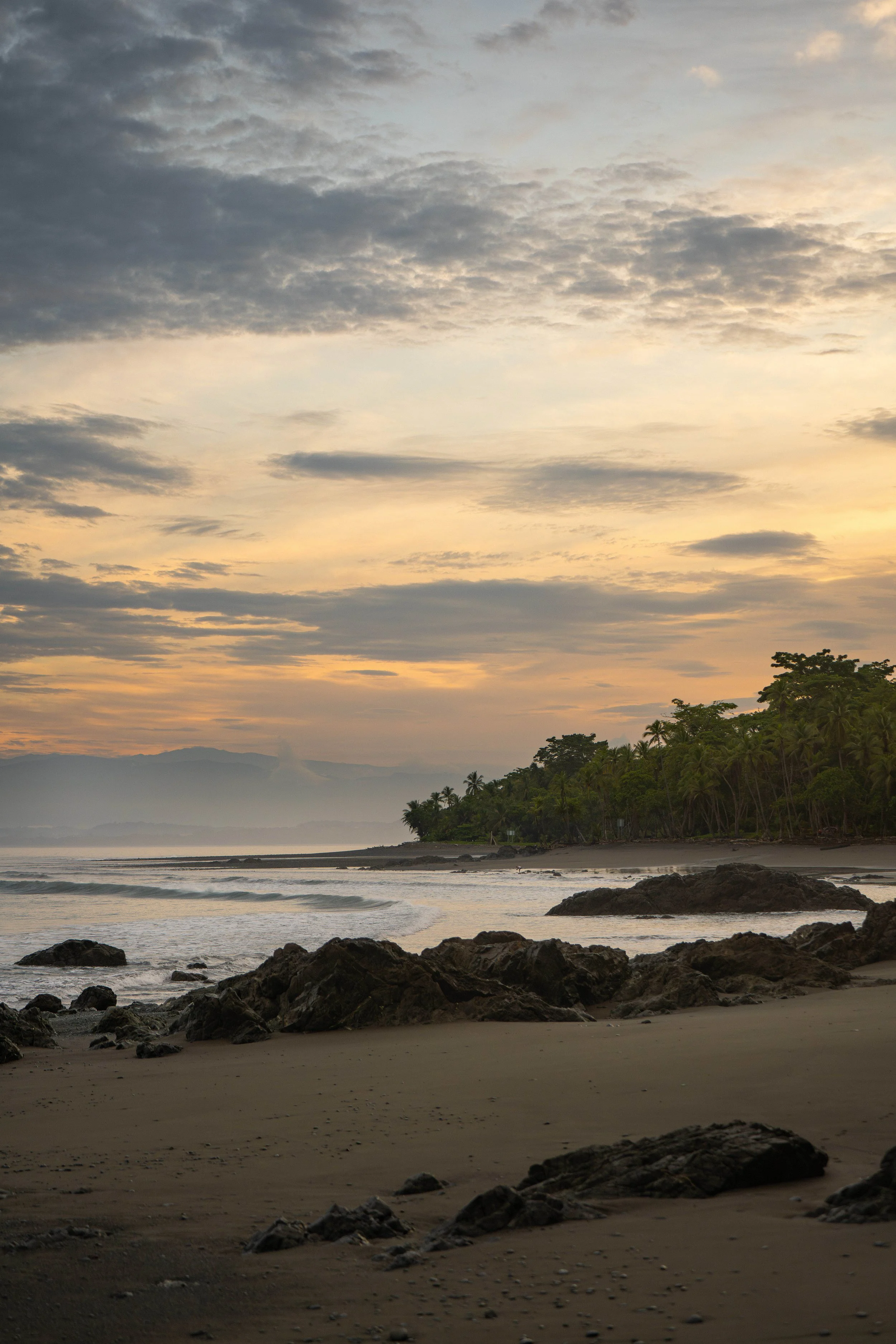 Sunset over a tropical beach with scattered rocks, lush green trees along the shoreline, and mountains in the background under a cloudy sky.
