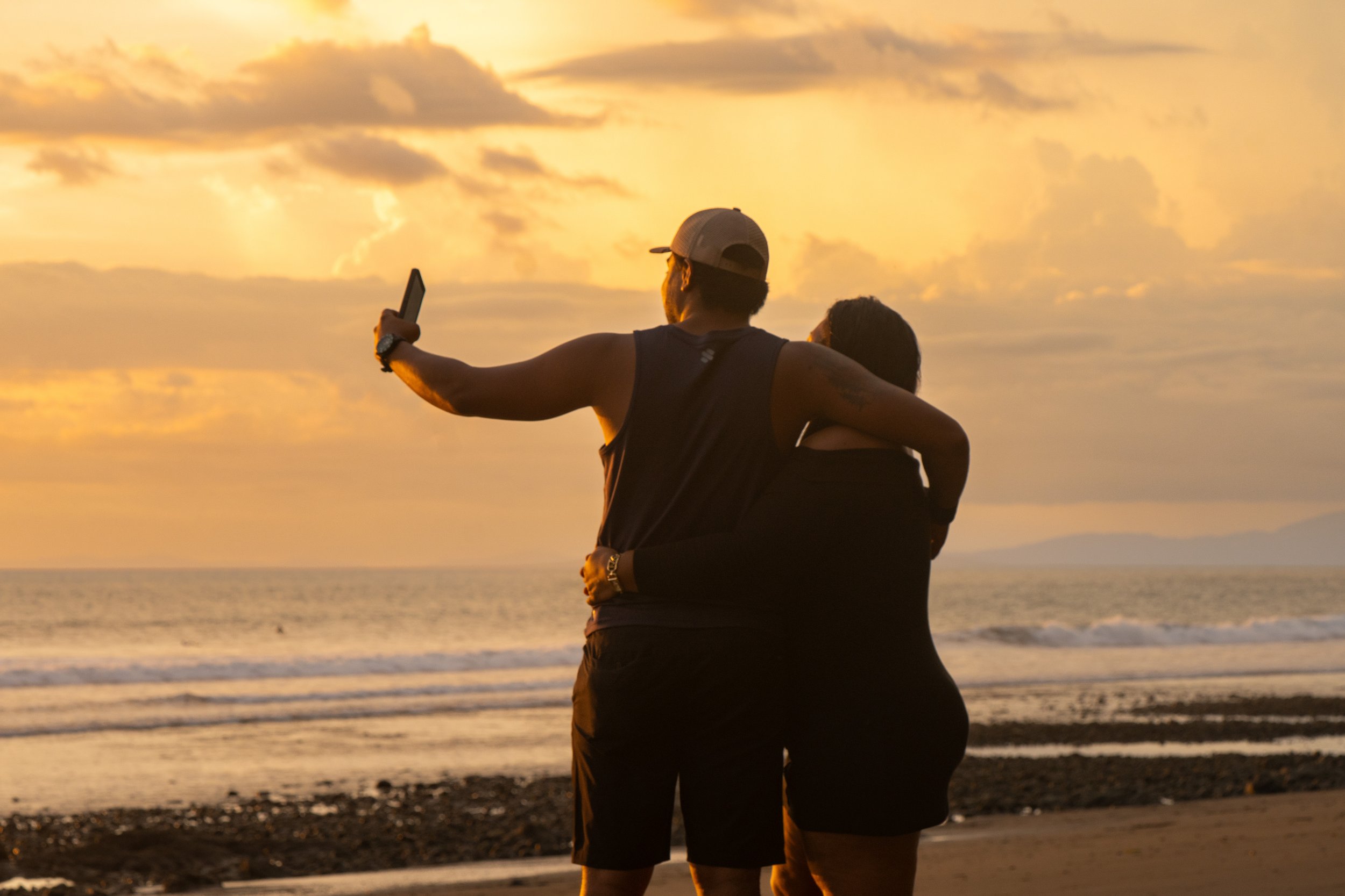 A man and woman standing on a beach during sunset, with the man taking a selfie using his phone.