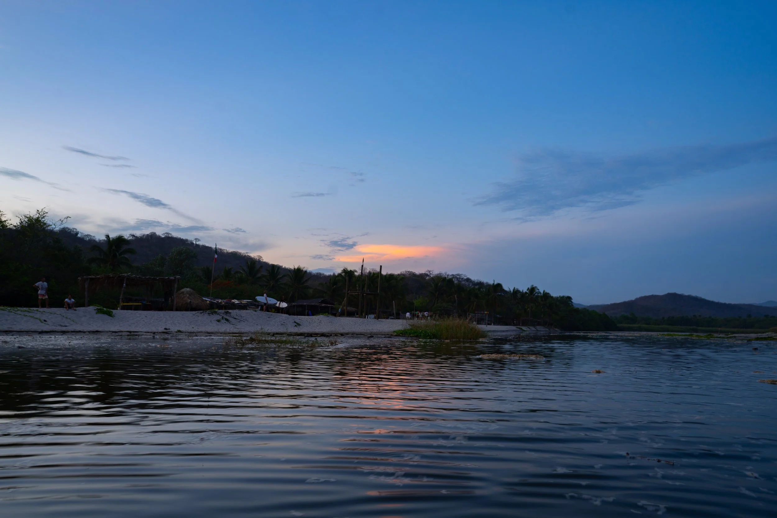 Scenic view of a river at dusk with a small village or settlement along the shoreline, surrounded by trees and hills, under a mostly clear sky with some clouds and a hint of sunset.