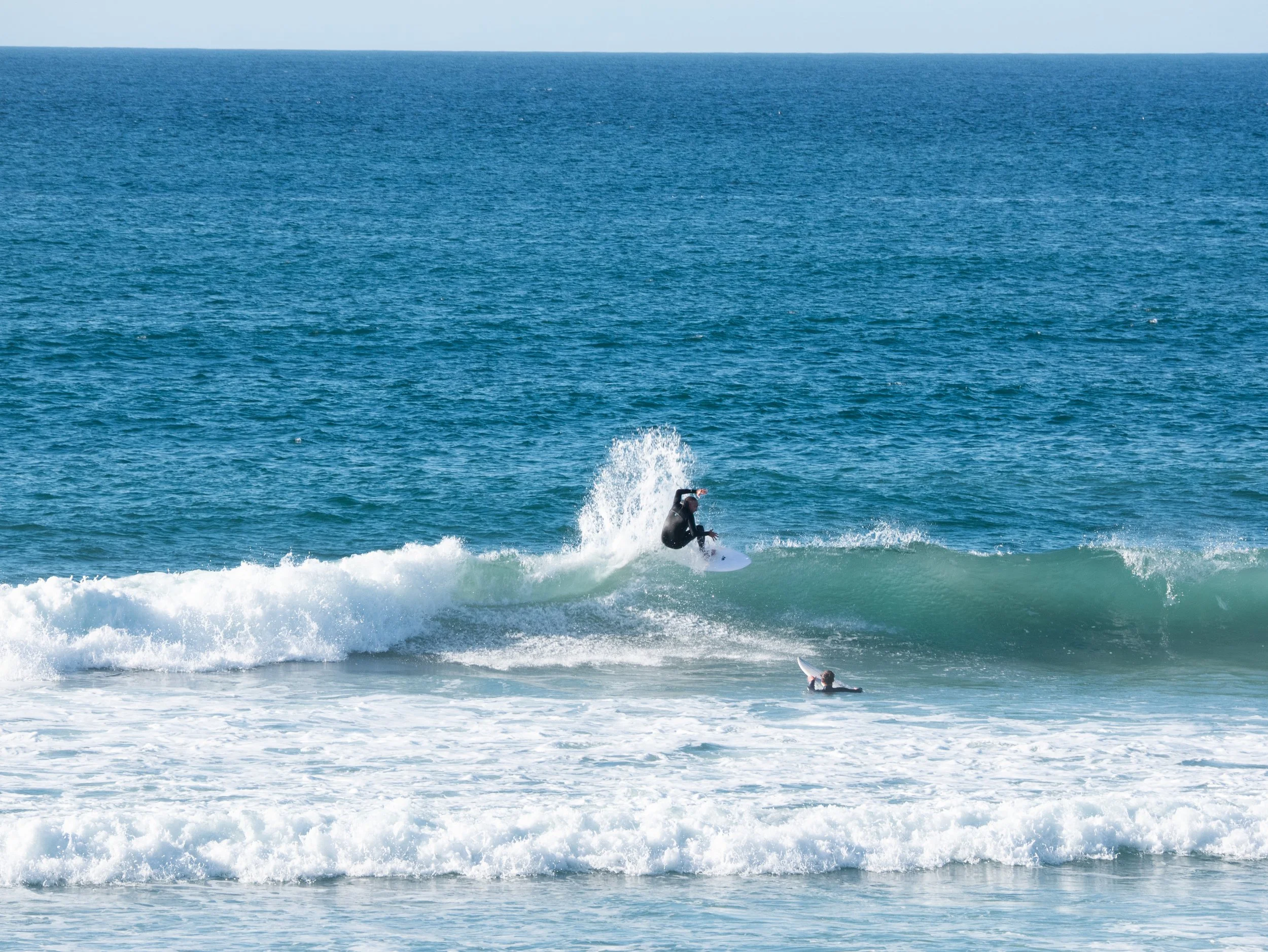 A person in a wetsuit surfing a wave in the ocean while another person floats nearby on a surfboard.